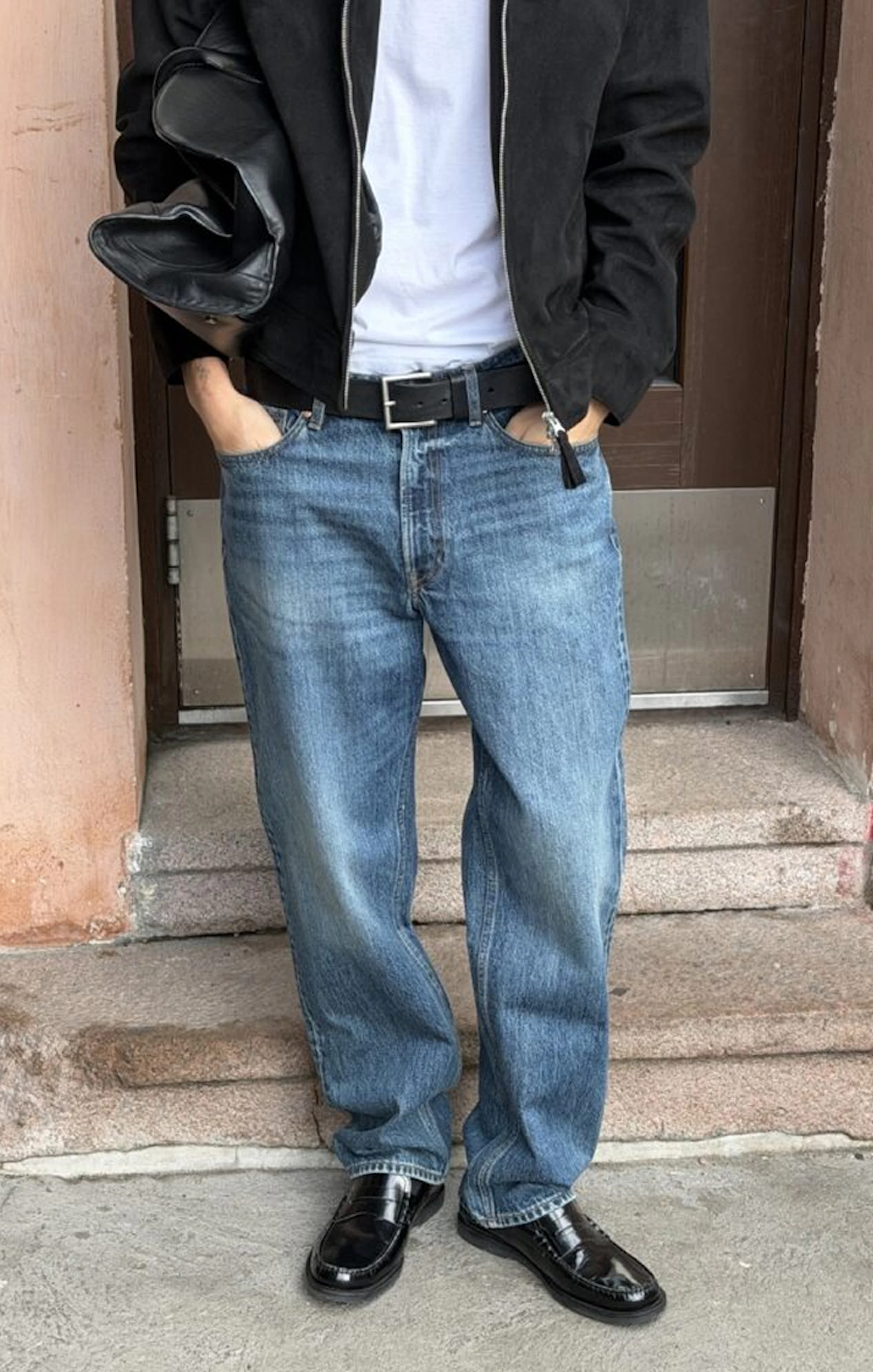 Person i svart jacka, vit t-shirt, blå jeans med svart bälte och svarta loafers, står framför en dörr.