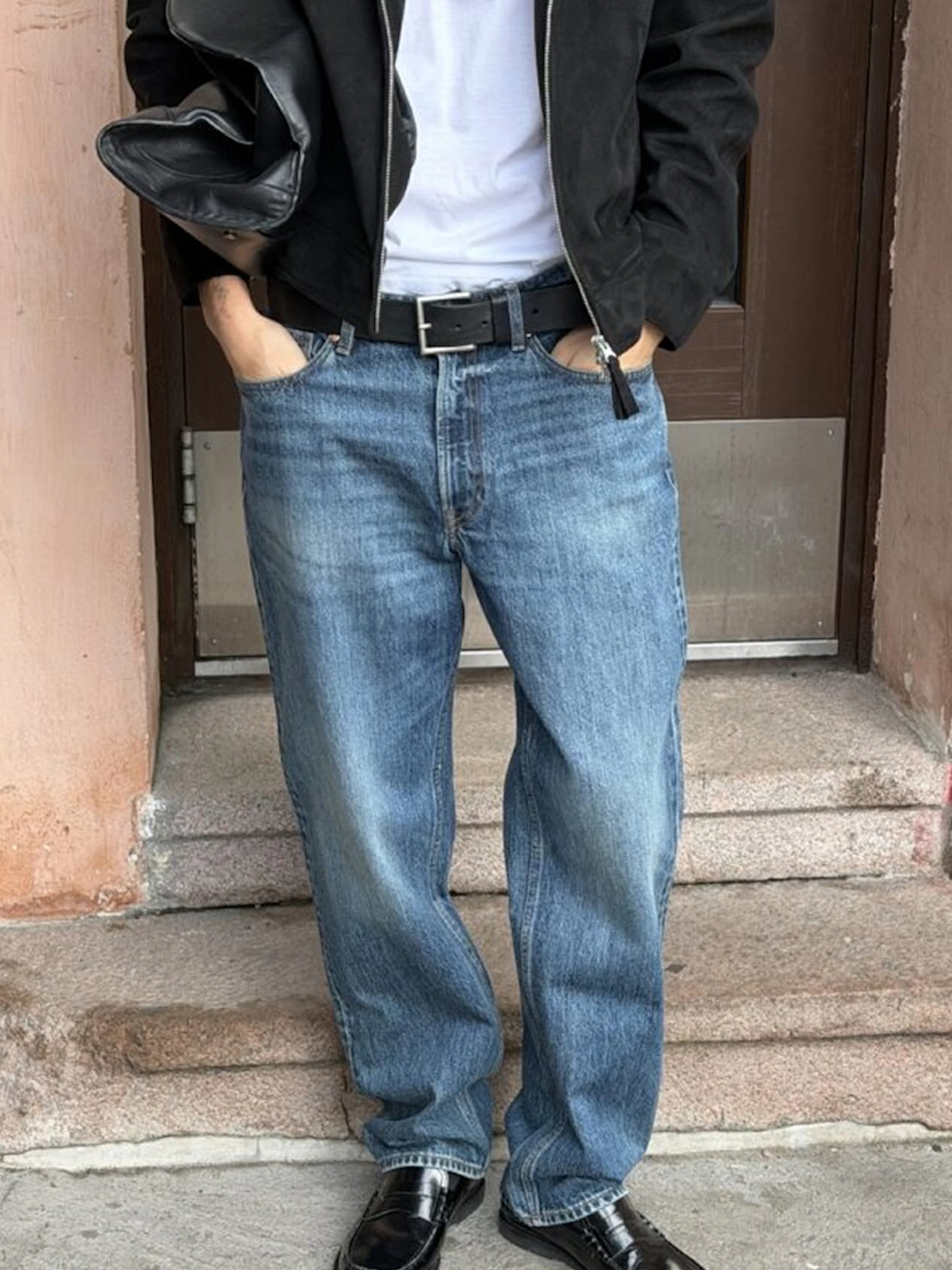 Person i svart jacka, vit t-shirt, blå jeans med svart bälte och svarta loafers, står framför en dörr.