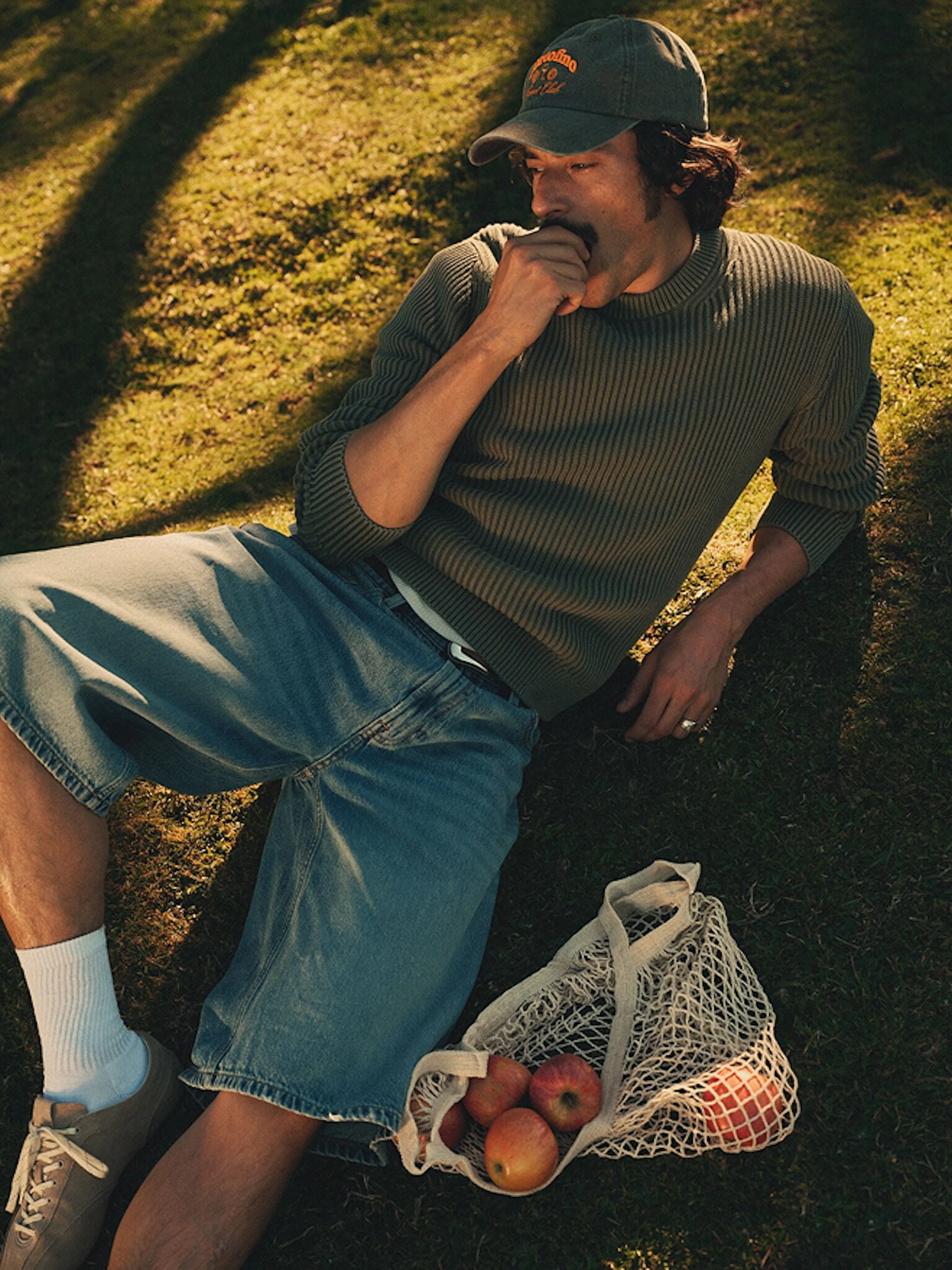 Young man in a green ribbed sweater and cap lying on grass, chewing his thumb, with a mesh bag of apples beside him.