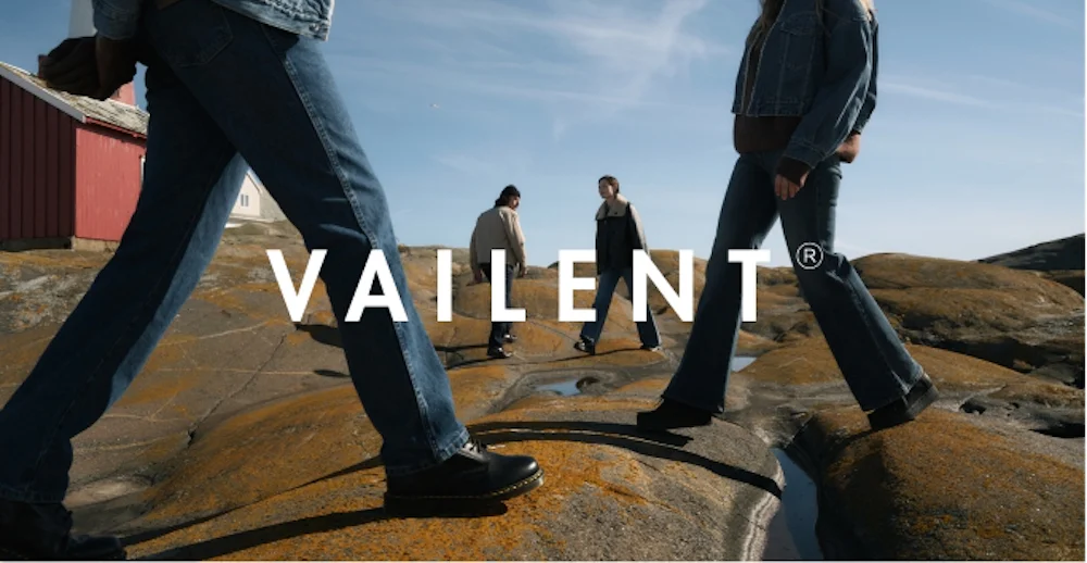 Fire personer i jeans går på fjellknauser ved kysten. Vailent-logo i midten. Rød hytte og blå himmel i bakgrunnen.