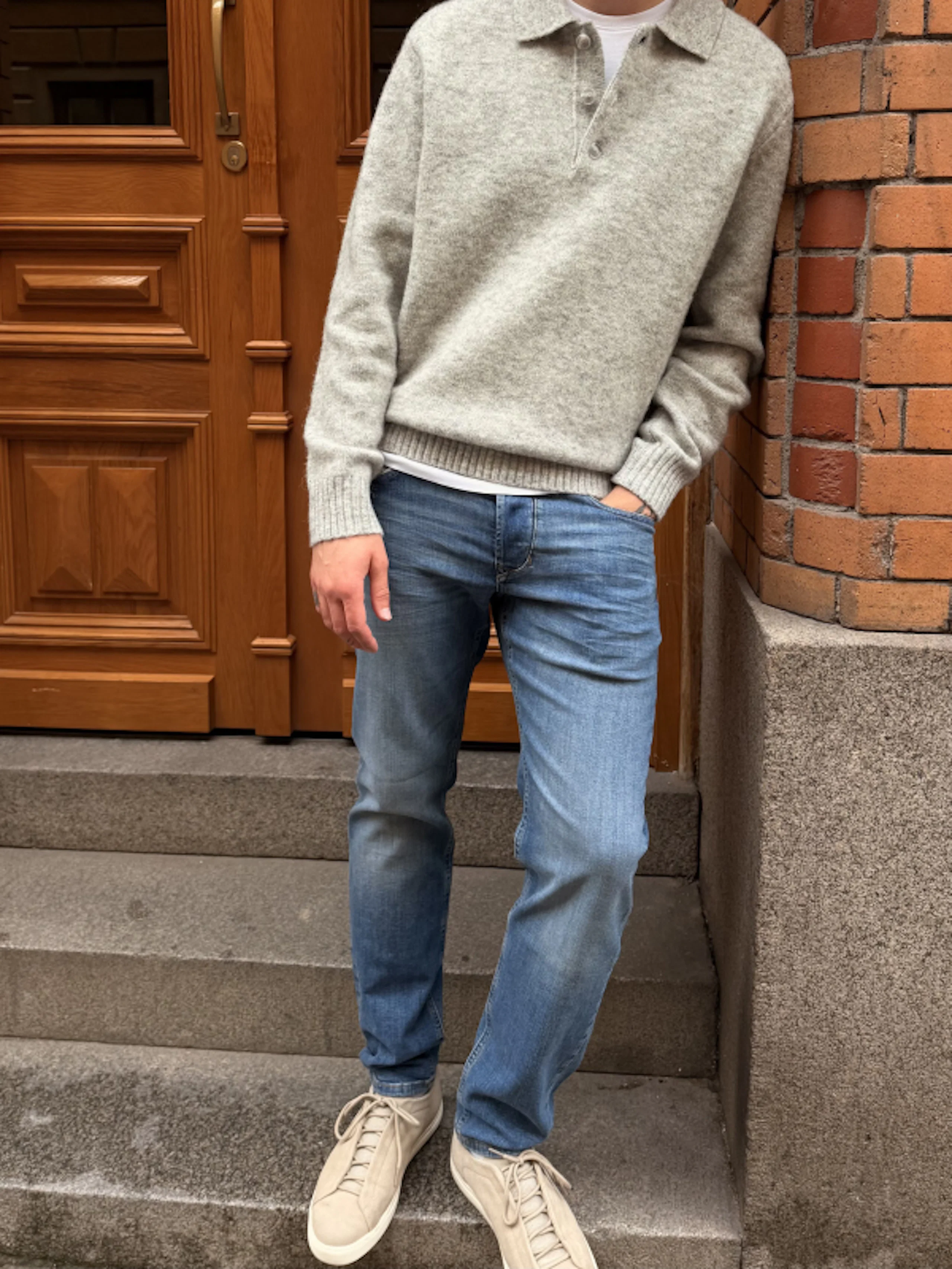 Mann iført grå genser, blå jeans og beige sneakers står på steintrapp, lent mot en murvegg ved siden av en tredør.