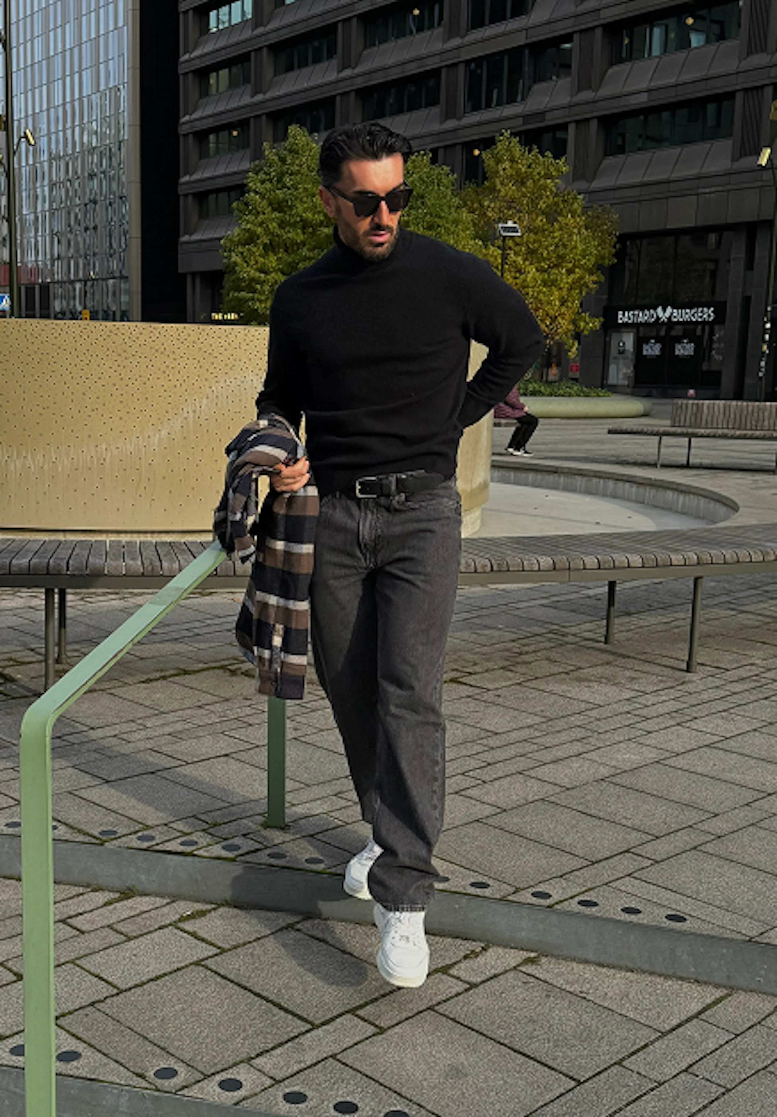 Mann i svart rullekragegenser, grå jeans og hvite sneakers går utendørs i byen, med en rutete jakke over armen.