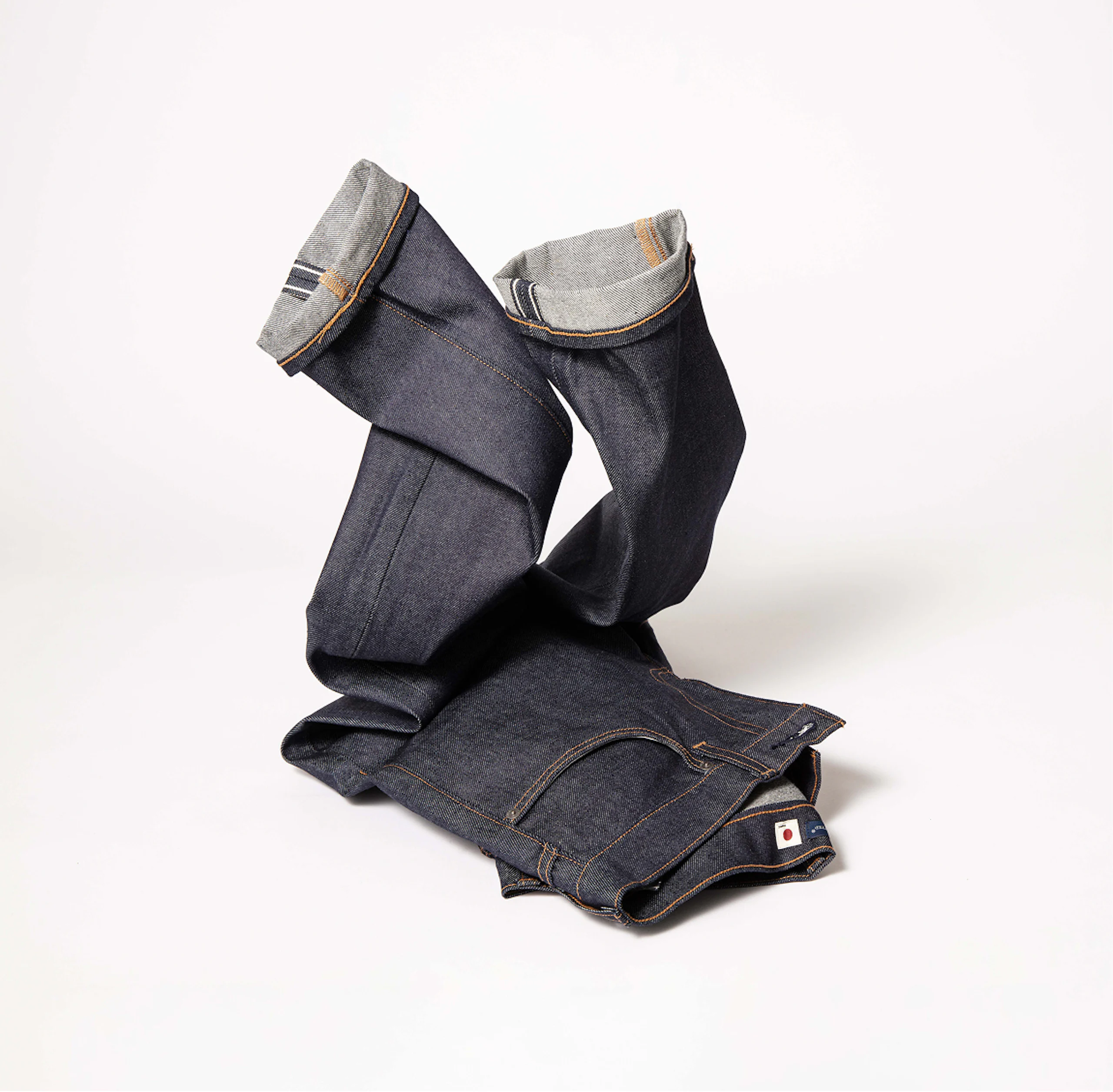 Mørk indigo rå denimjeans, brettet med opprullede mansjetter som viser selvedge og oransje søm, lagt på hvit bakgrunn.