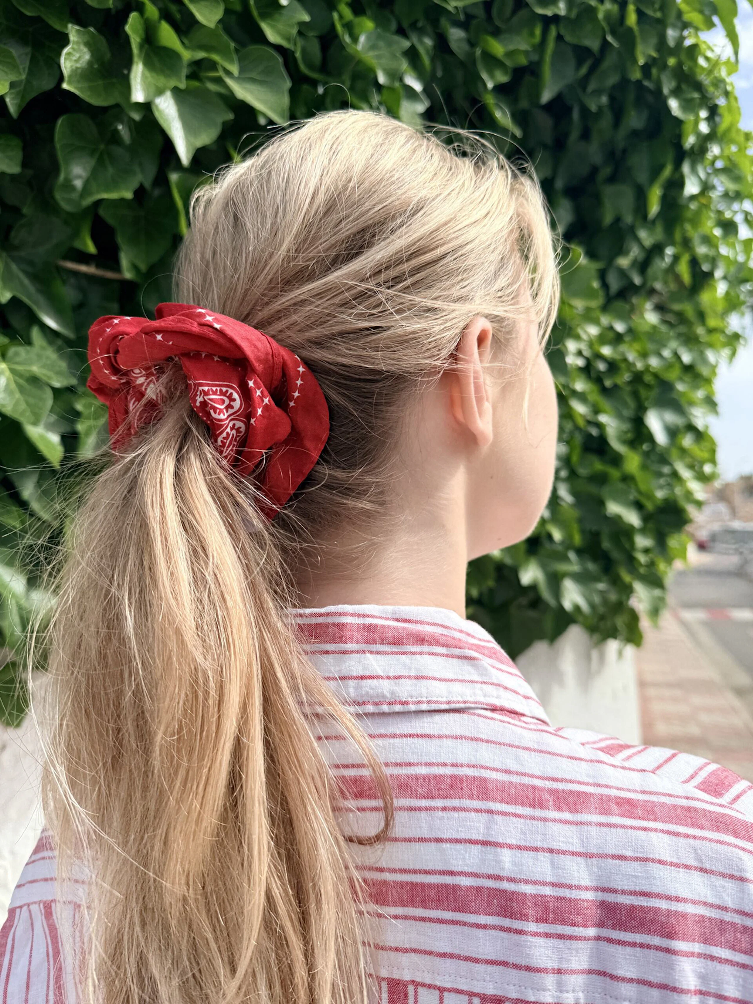 Kvinna med blont hår i hästsvans, fäst med röd scrunchie med paisleymönster. Randig röd-vit topp, grön murgröna i bakgrunden.