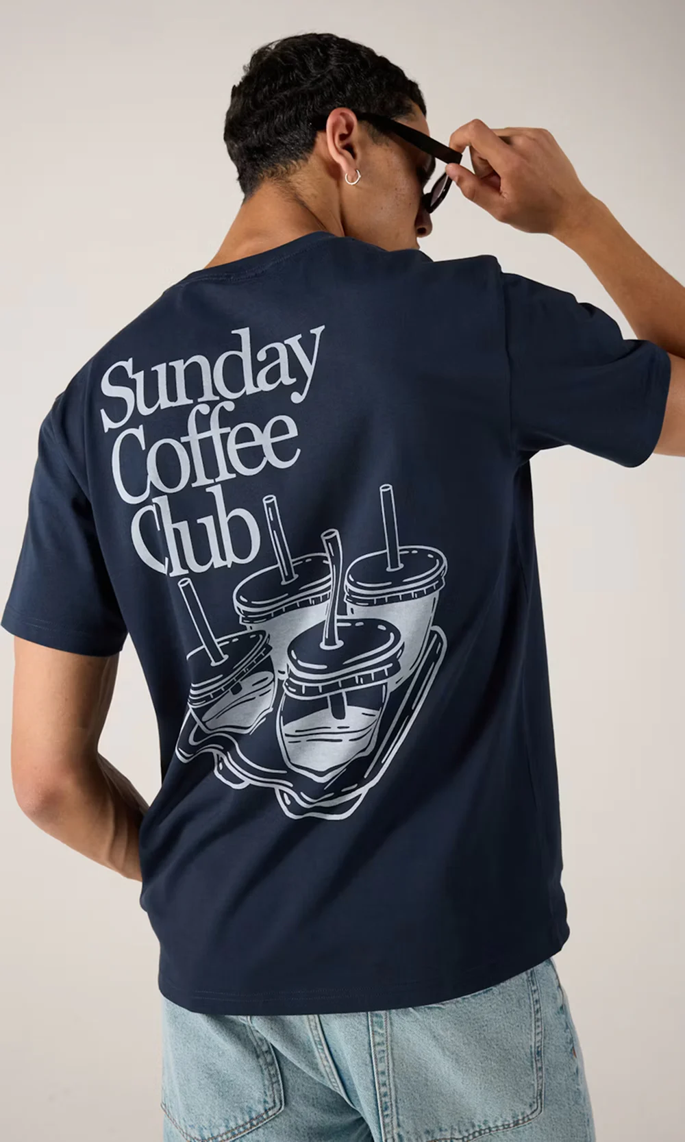 Man bakifrån i marinblå t-shirt med "Sunday Coffee Club"-tryck och kaffekopp-illustration, ljusblå jeans.
