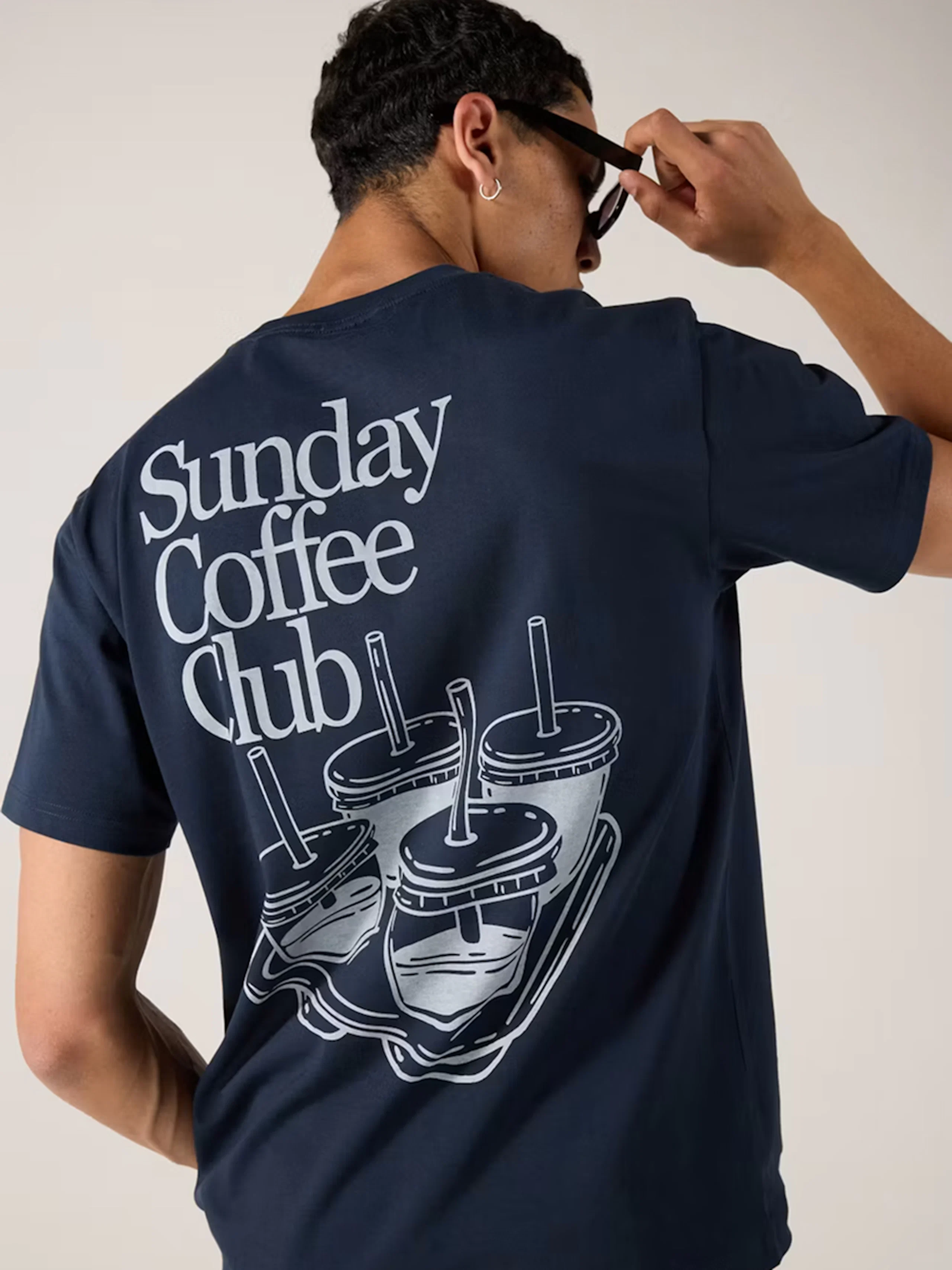 Man bakifrån i marinblå t-shirt med "Sunday Coffee Club"-tryck och kaffekopp-illustration, ljusblå jeans.