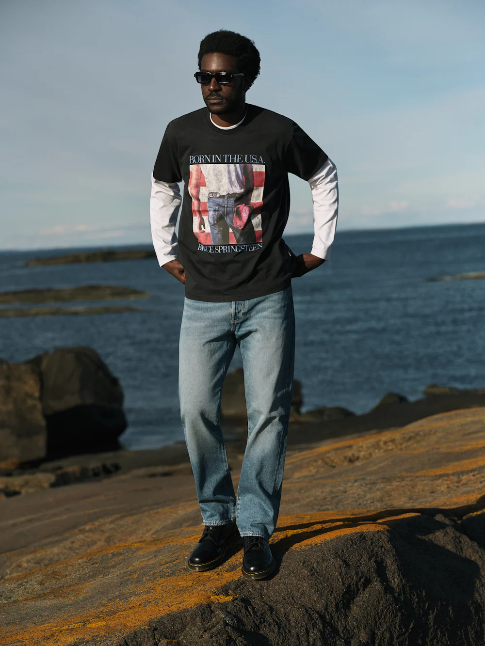Svart mann med solbriller, iført svart "Born in the U.S.A. Bruce Springsteen"-T‑skjorte over hvit langermet genser og lyseblå jeans, stående på steinete strand med havet i bakgrunnen.