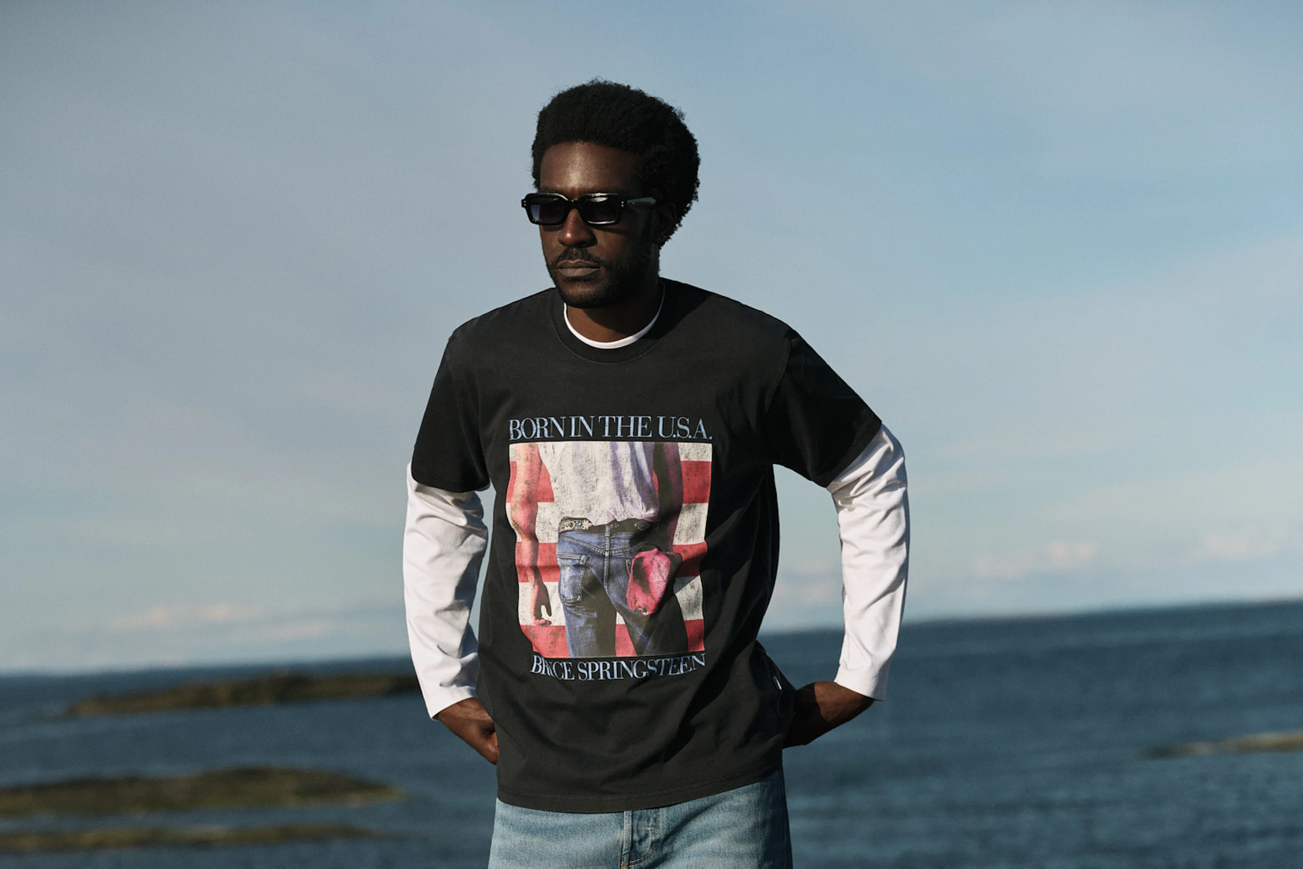 Svart mann med solbriller, iført svart "Born in the U.S.A. Bruce Springsteen"-T‑skjorte over hvit langermet genser og lyseblå jeans, stående på steinete strand med havet i bakgrunnen.