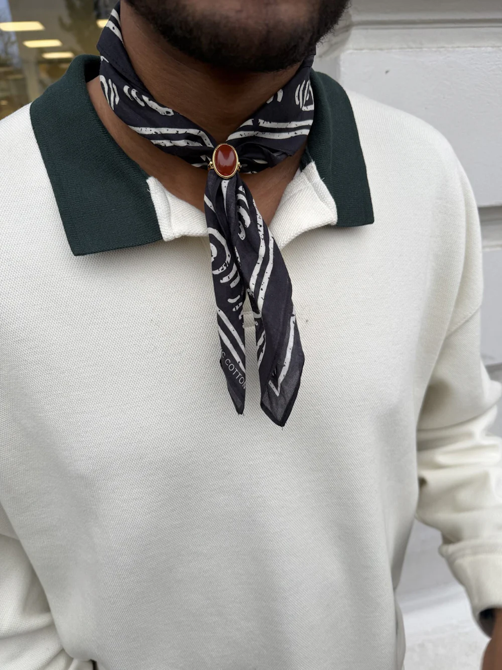 Man bär vit pikétröja med grön krage och ett mönstrat mörkgrått scarfhalsband fäst med en guldbrosch med röd sten.