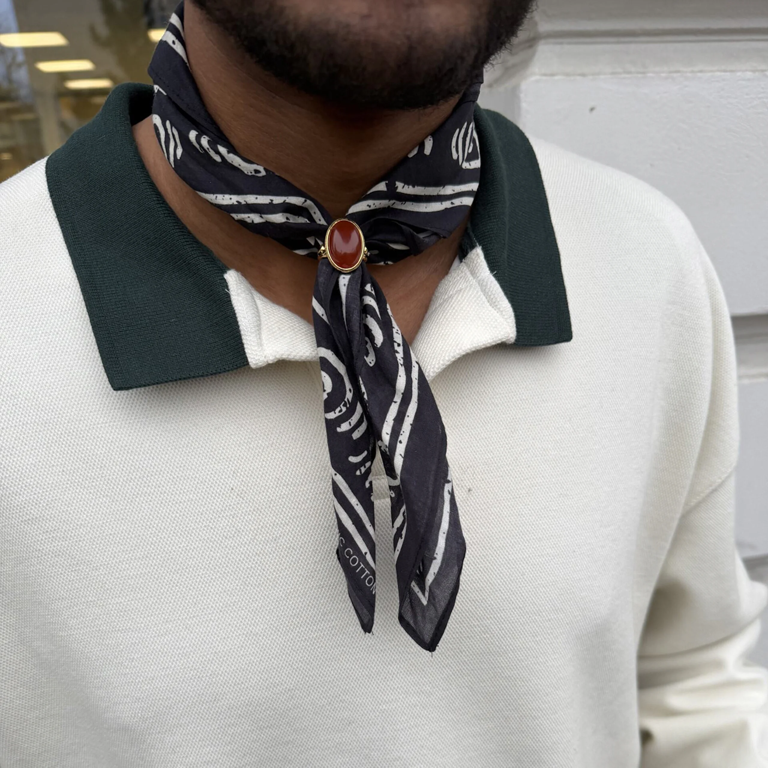 Man bär vit pikétröja med grön krage och ett mönstrat mörkgrått scarfhalsband fäst med en guldbrosch med röd sten.
