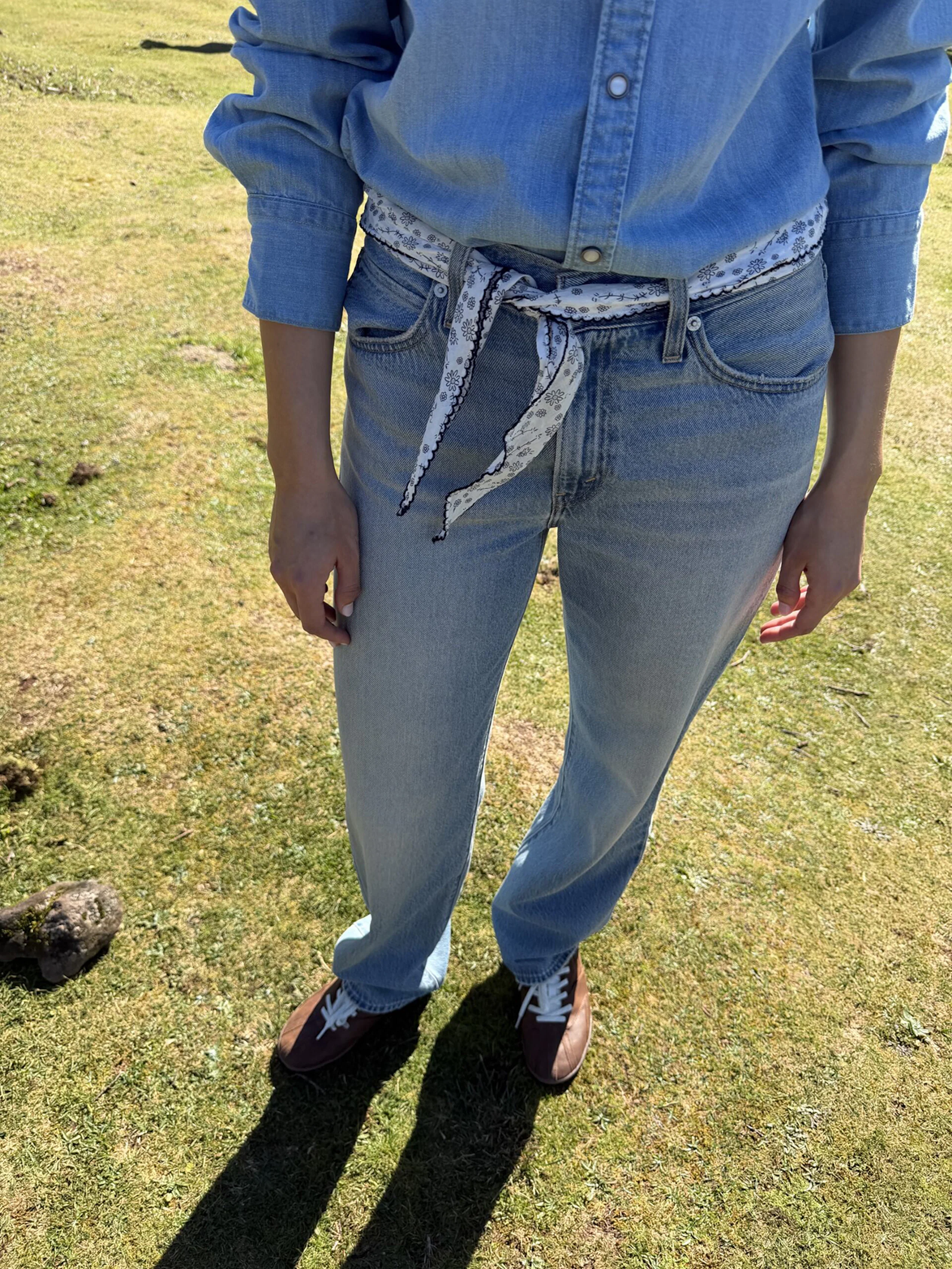 Person i ljusblå jeansskjorta och jeans med ett vitt mönstrat scarf knutet runt midjan, utomhus på gräs.