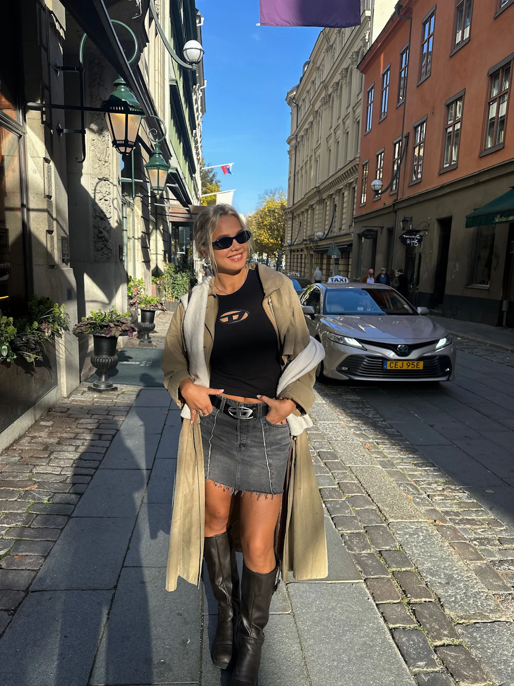Ung kvinne i sort topp, denimskjørt, lang beige frakk og sorte støvletter poserer smilende på en solrik bygate.