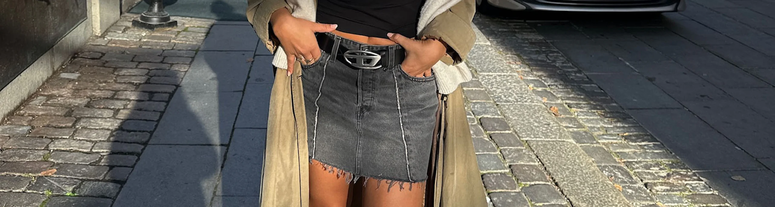 Ung kvinne i sort topp, denimskjørt, lang beige frakk og sorte støvletter poserer smilende på en solrik bygate.