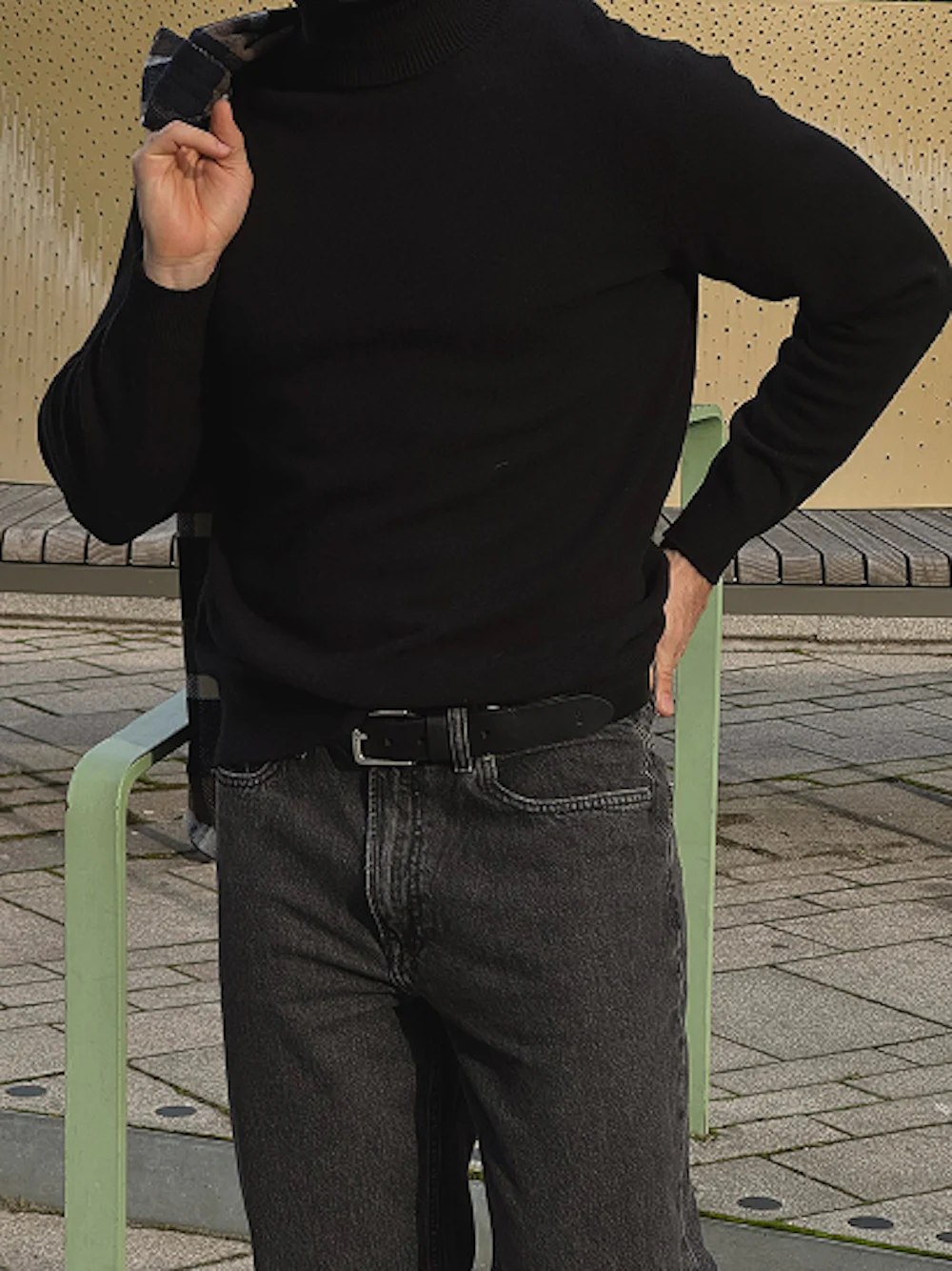 Person i svart genser og mørke jeans med svart belte, stående utendørs ved en lysegrønn benk.