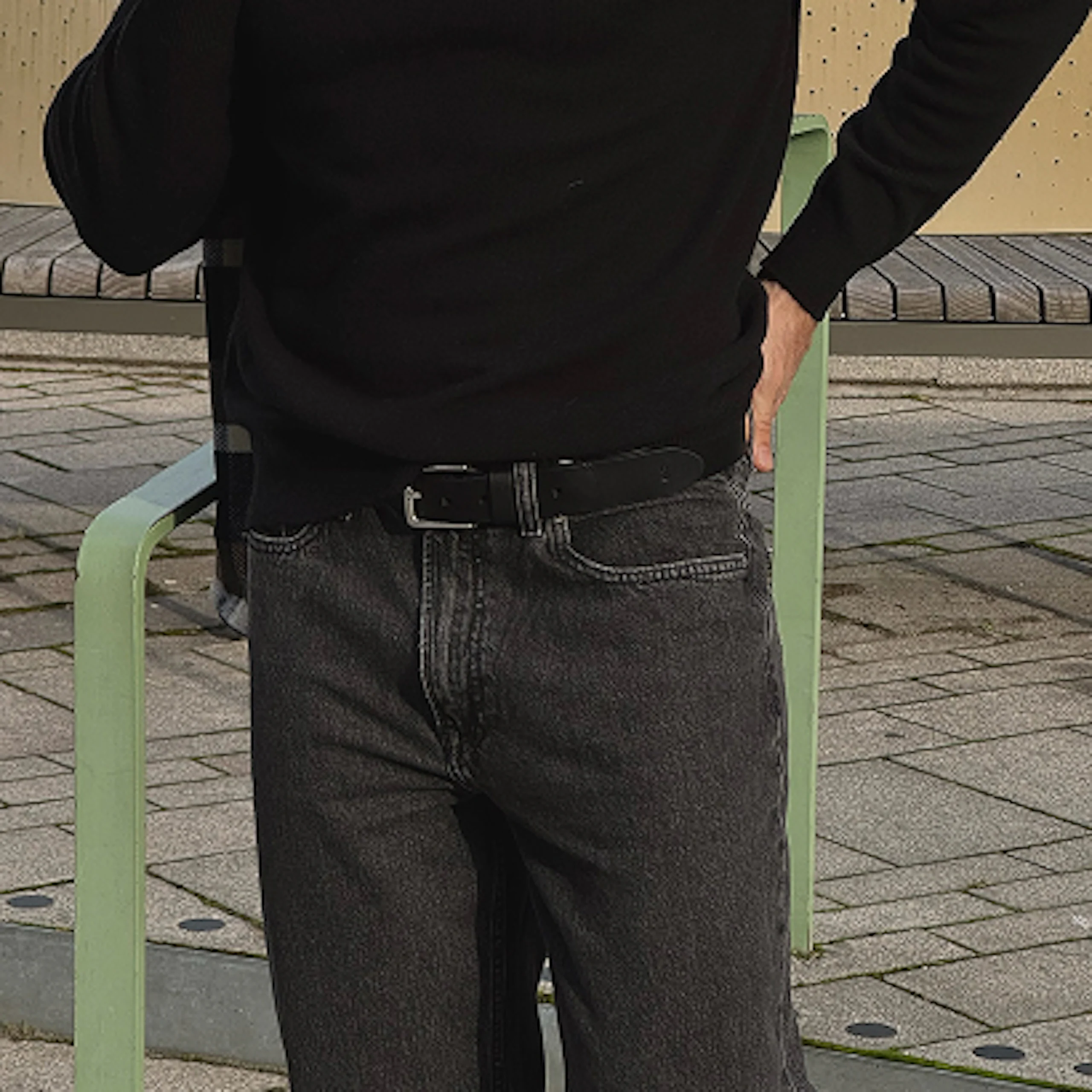 Person i svart genser og mørke jeans med svart belte, stående utendørs ved en lysegrønn benk.