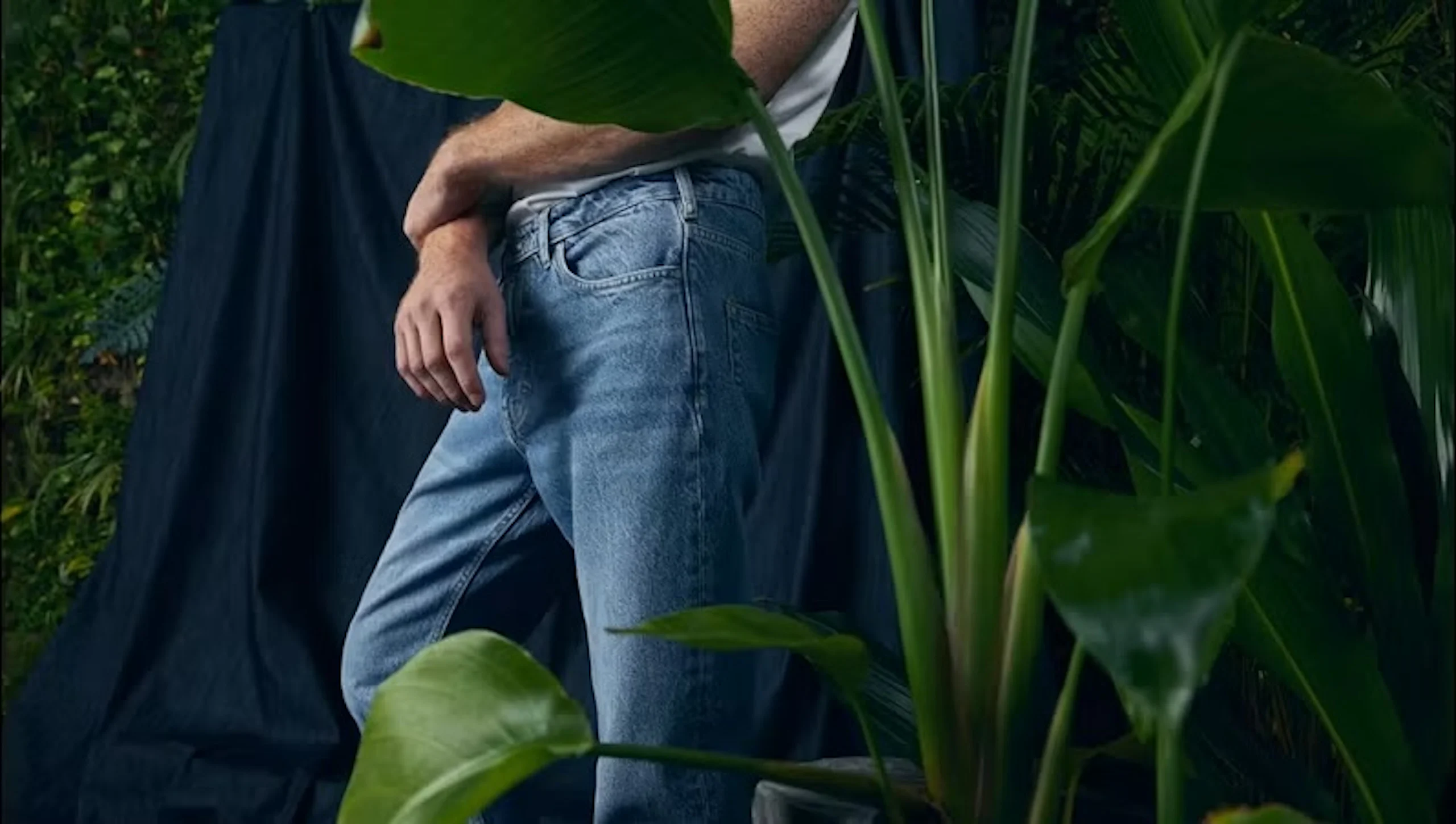 Närbild på en person som bär ljusblå jeans och en vit skjorta, stående med händerna nära fickorna bland stora gröna tropiska krukväxtblad.