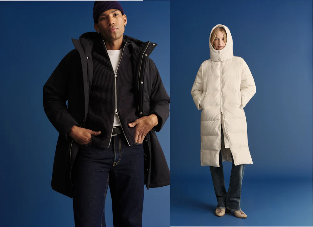 Man wearing black parka and jeans with hands in pockets beside woman in long white puffer coat with hood up against blue backdrop.