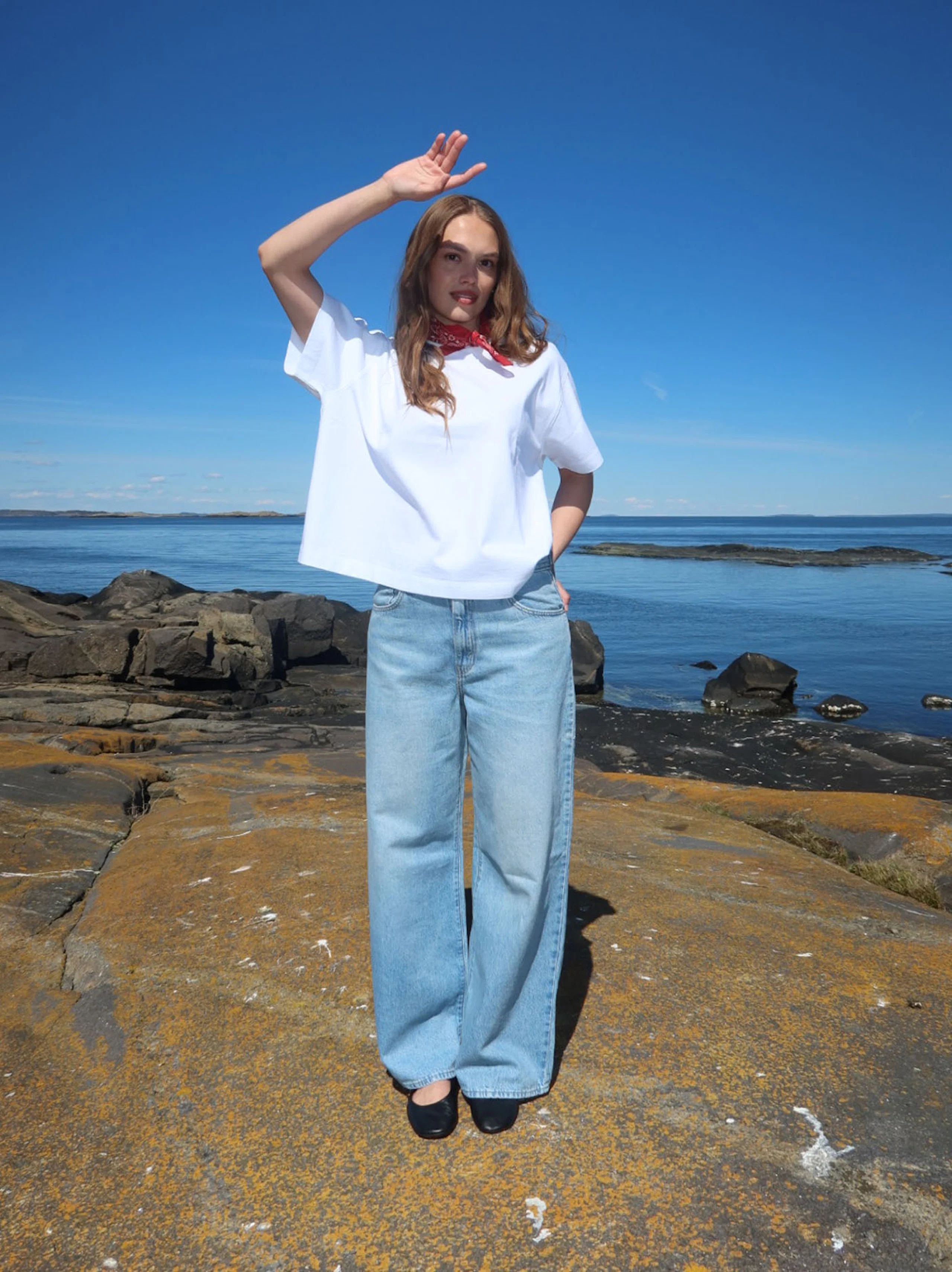 Kvinne i hvit t-skjorte, lyse wide-leg jeans og rød bandana står på svaberg ved havet under blå himmel.