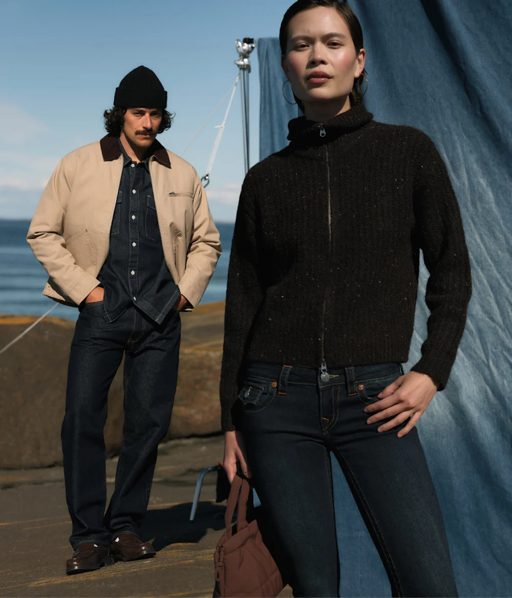 To personer i casual høstantrekk ved en havn. Mann i beige jakke og lue, kvinne i mørk strikkejakke og jeans.