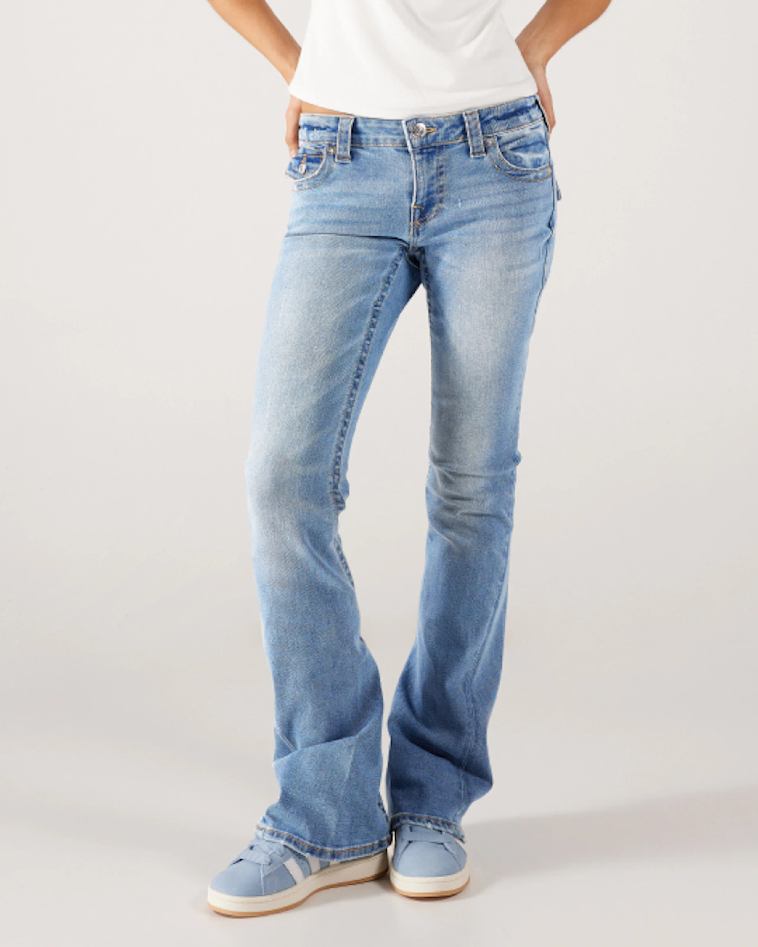 Kvinna i ljusblå utsvängda jeans med låg midja, vit topp och blå sneakers mot grå bakgrund.