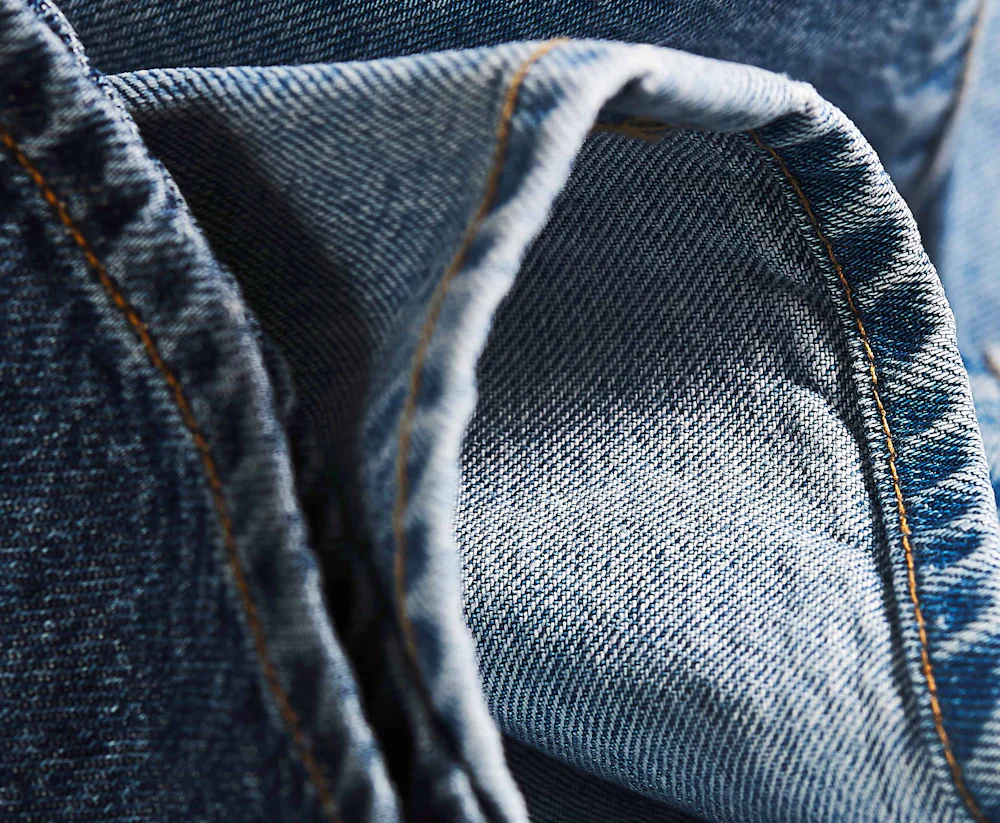 Nærbilde av kanten på en blå denimjeans som fremhever den karakteristiske denimstrukturen, slitesterke kontrastsømmer og stoffets kvalitet.