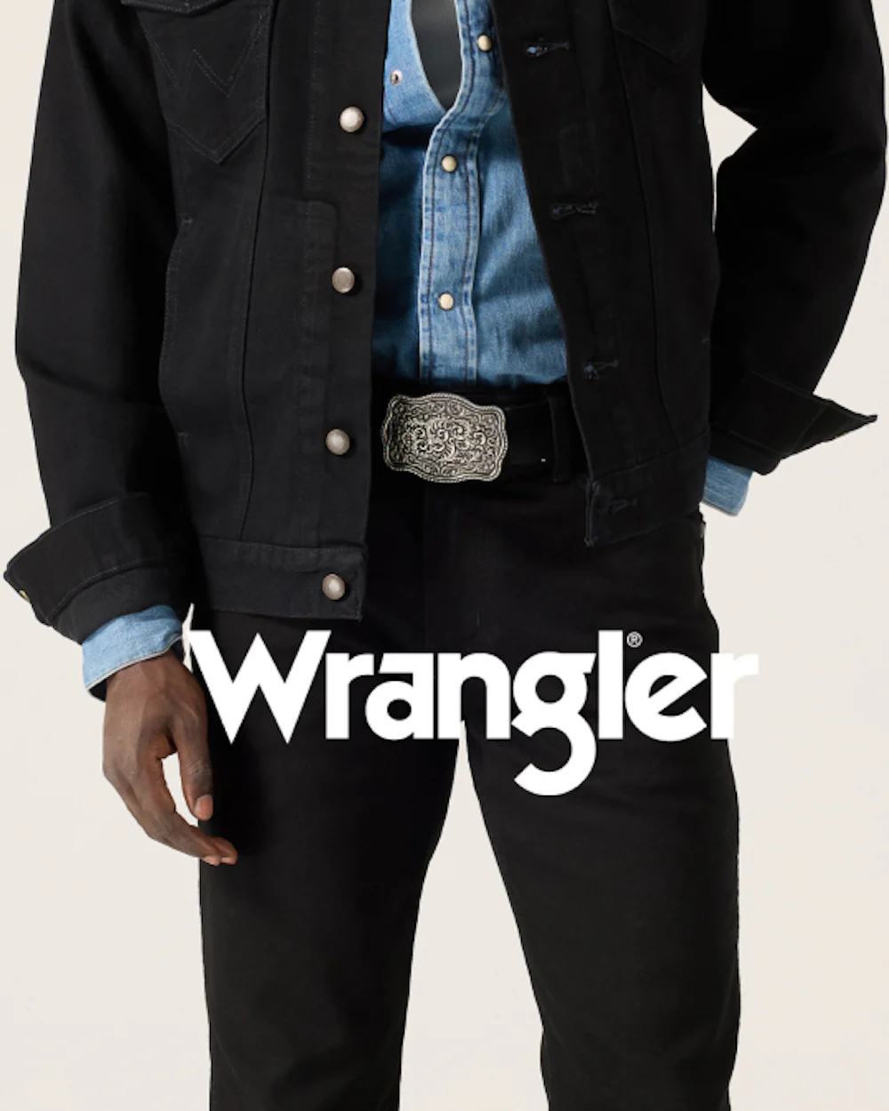 Mann iført svart Wrangler denimjakke over blå skjorte med dekorativt beltespenne og svarte jeans. Wrangler-logo vises.