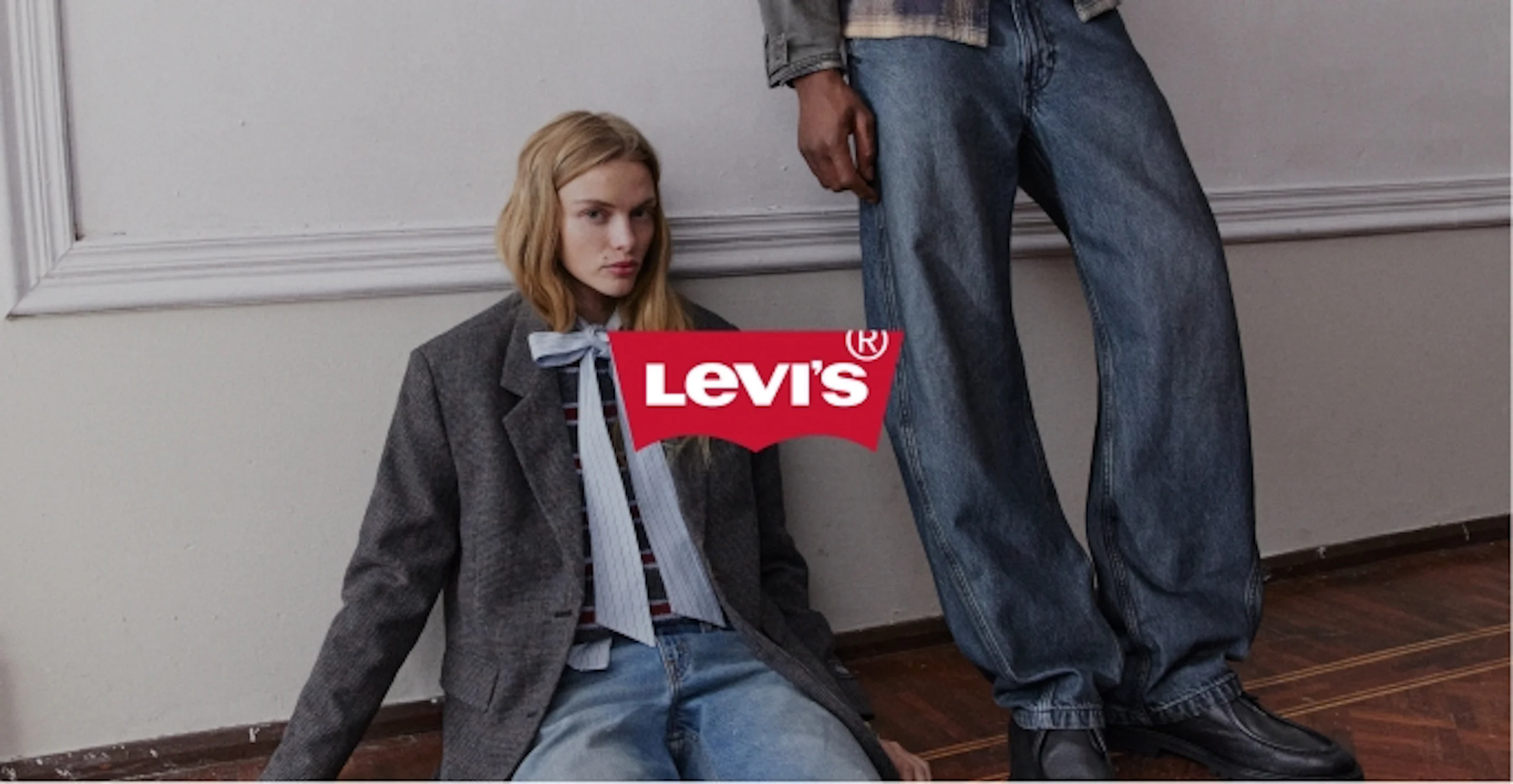 Nainen istuu lattialla harmaassa takissa ja farkuissa, mies seisoo vieressä leveissä farkuissa. Levi's-logo kuvassa.