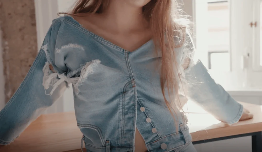 Nærbilde av en person som har på seg en slitt lyseblå denimjakke med frynsete hull på ermene og knappedetaljer, armene utstrakt i et solfylt kjøkkenmiljø.