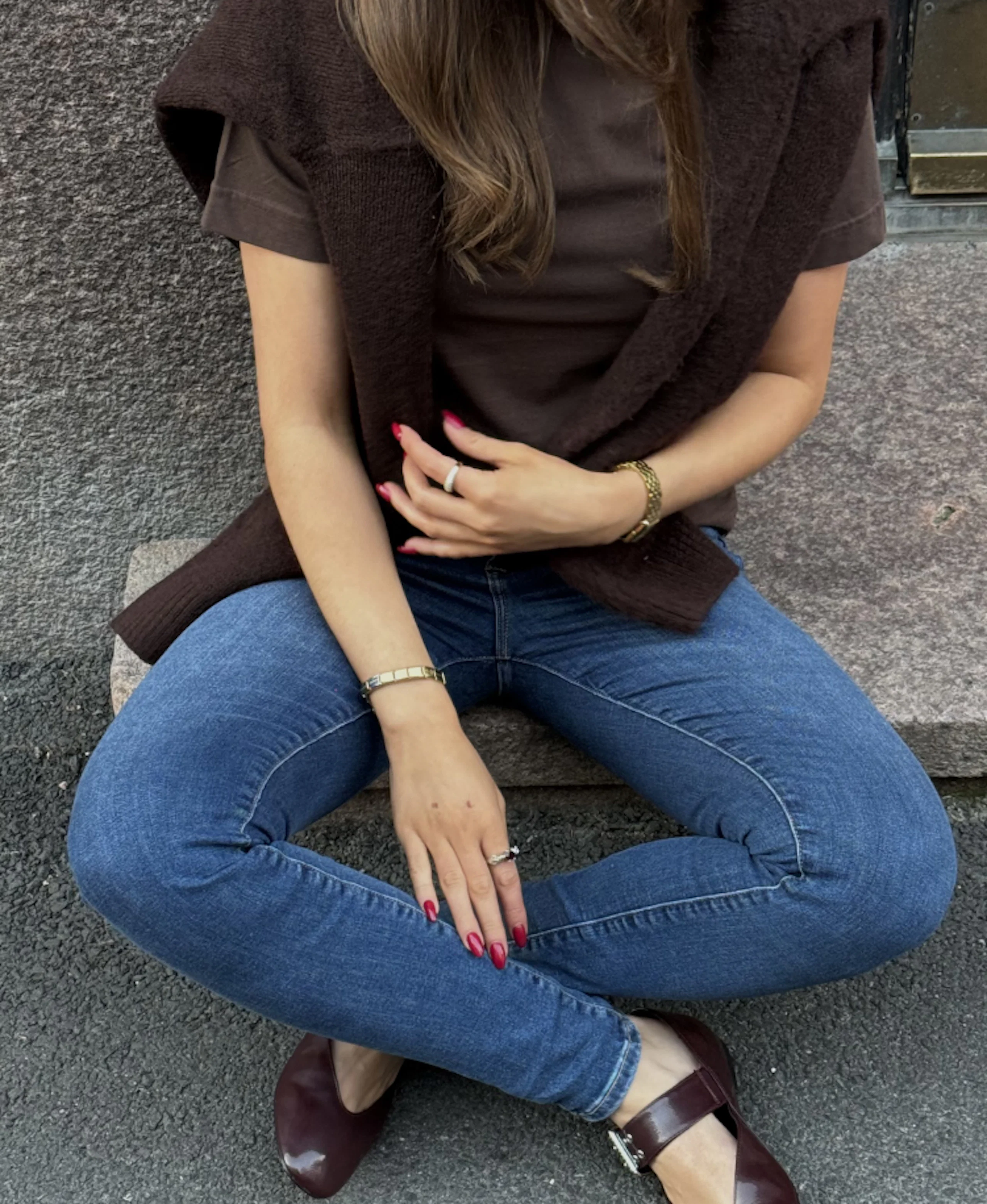 Kvinna sitter på trappa i blå jeans, brun tröja och mörkröda skor. Röda naglar, guldarmbånd och ringar syns tydligt.