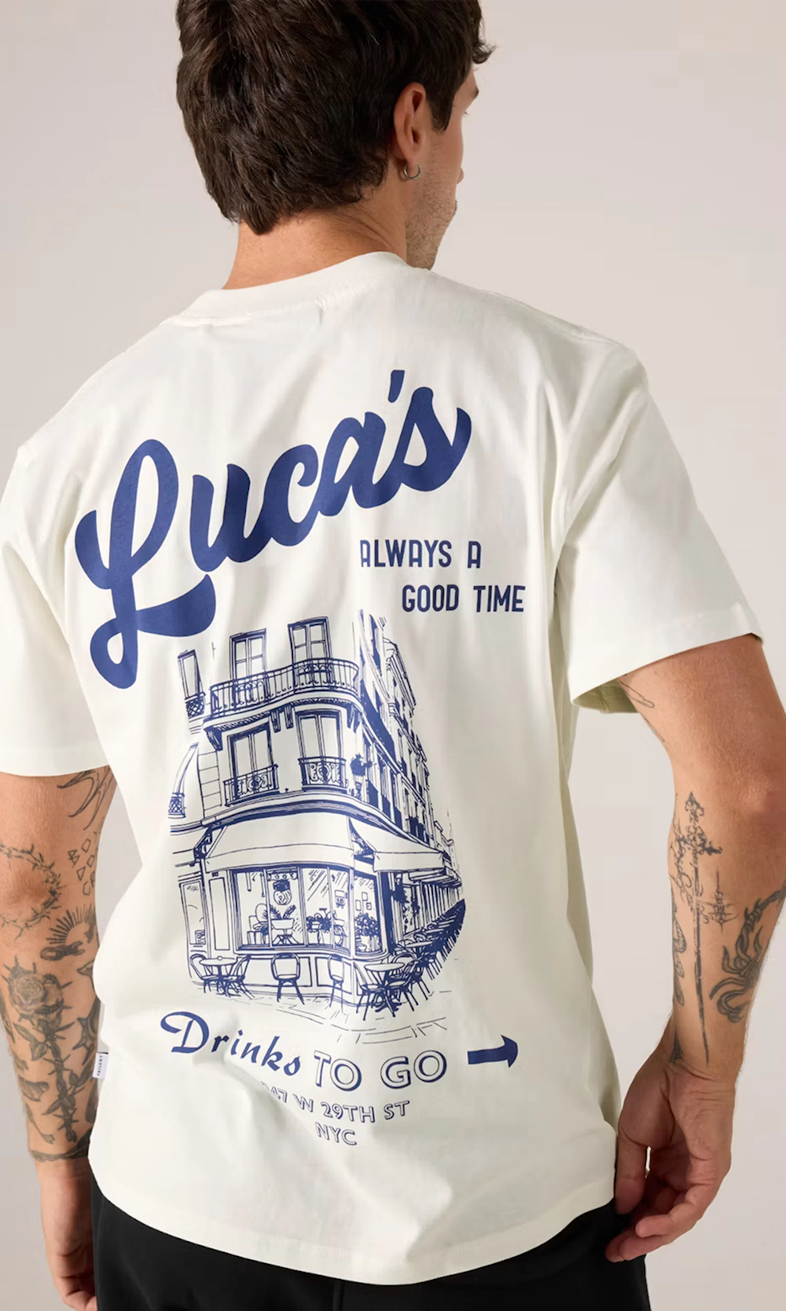 Man bakifrån i vit t-shirt med blått tryck: "Luca's Always a Good Time" och illustration av NYC-restaurang. Tatueringar på armarna.