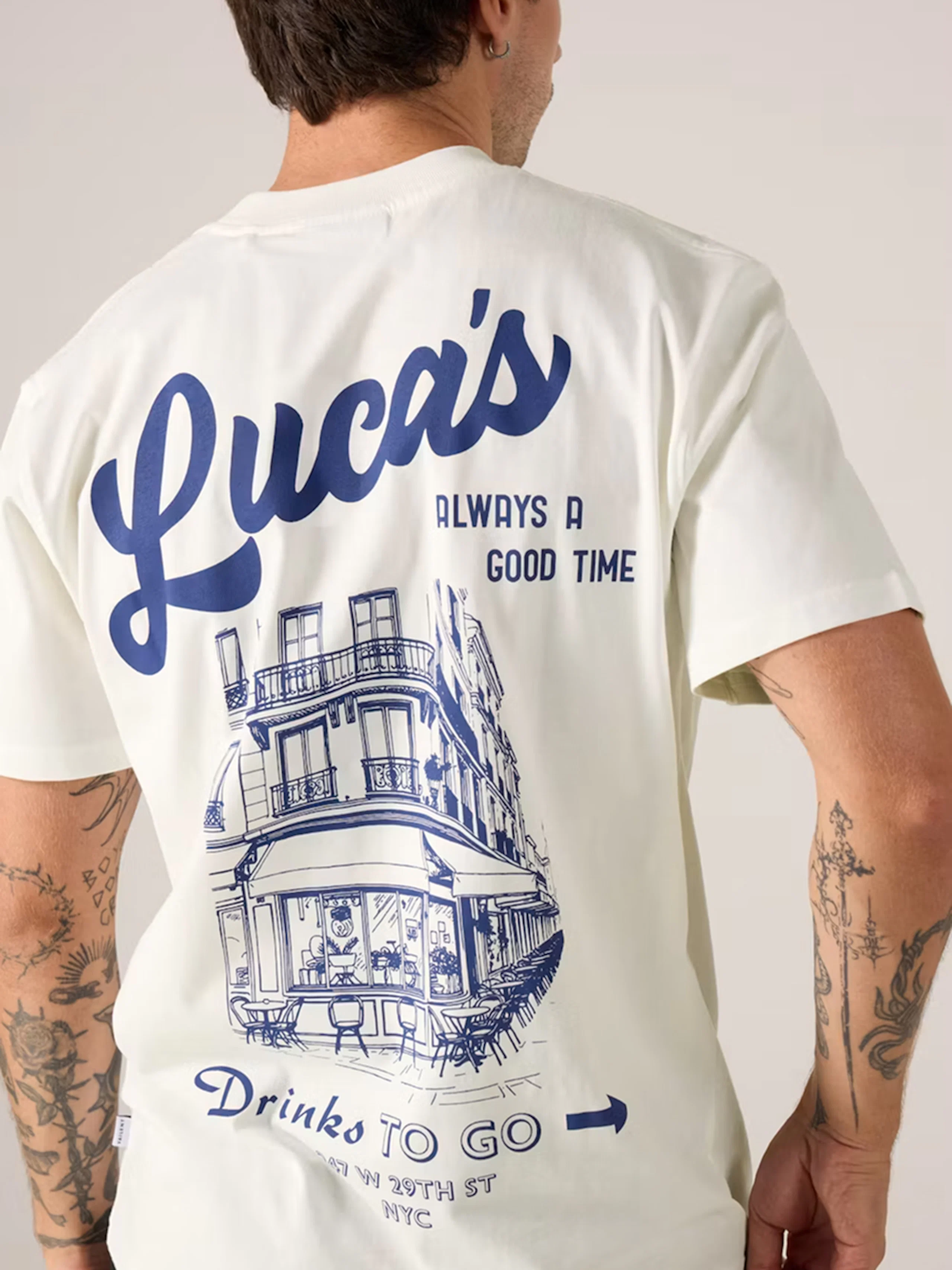 Man bakifrån i vit t-shirt med blått tryck: "Luca's Always a Good Time" och illustration av NYC-restaurang. Tatueringar på armarna.