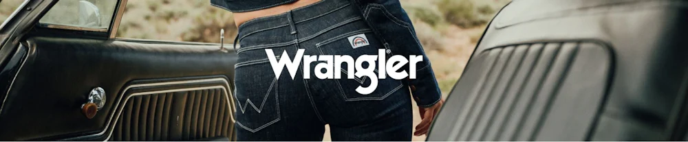 Henkilö tummansinisessä Wrangler-farkkuissa vanhan auton vieressä. Wrangler-logo kuvassa.