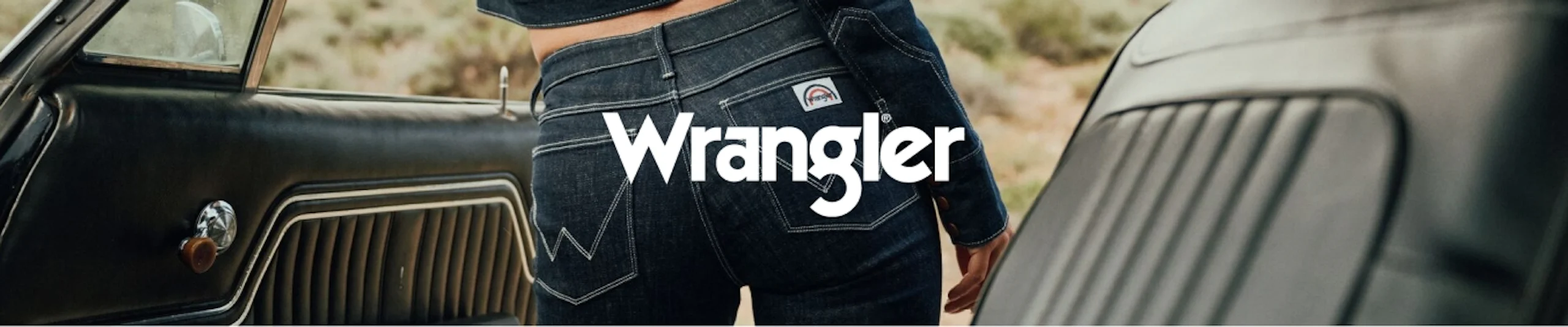 Henkilö tummansinisessä Wrangler-farkkuissa vanhan auton vieressä. Wrangler-logo kuvassa.