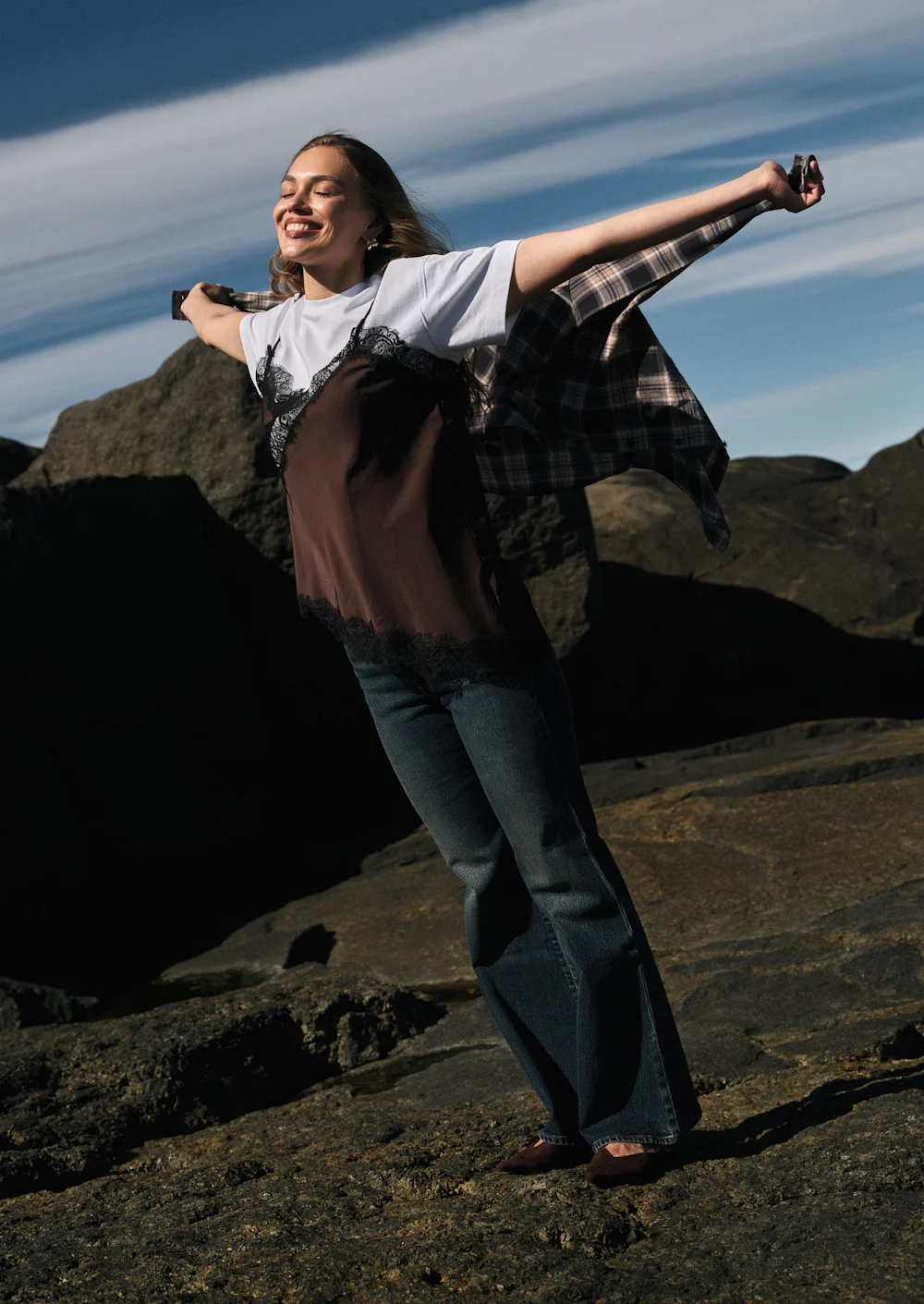 Woman smiling with arms outstretched on rocky terrain, wearing jeans, white tee, and plaid shirt, under a blue sky.
