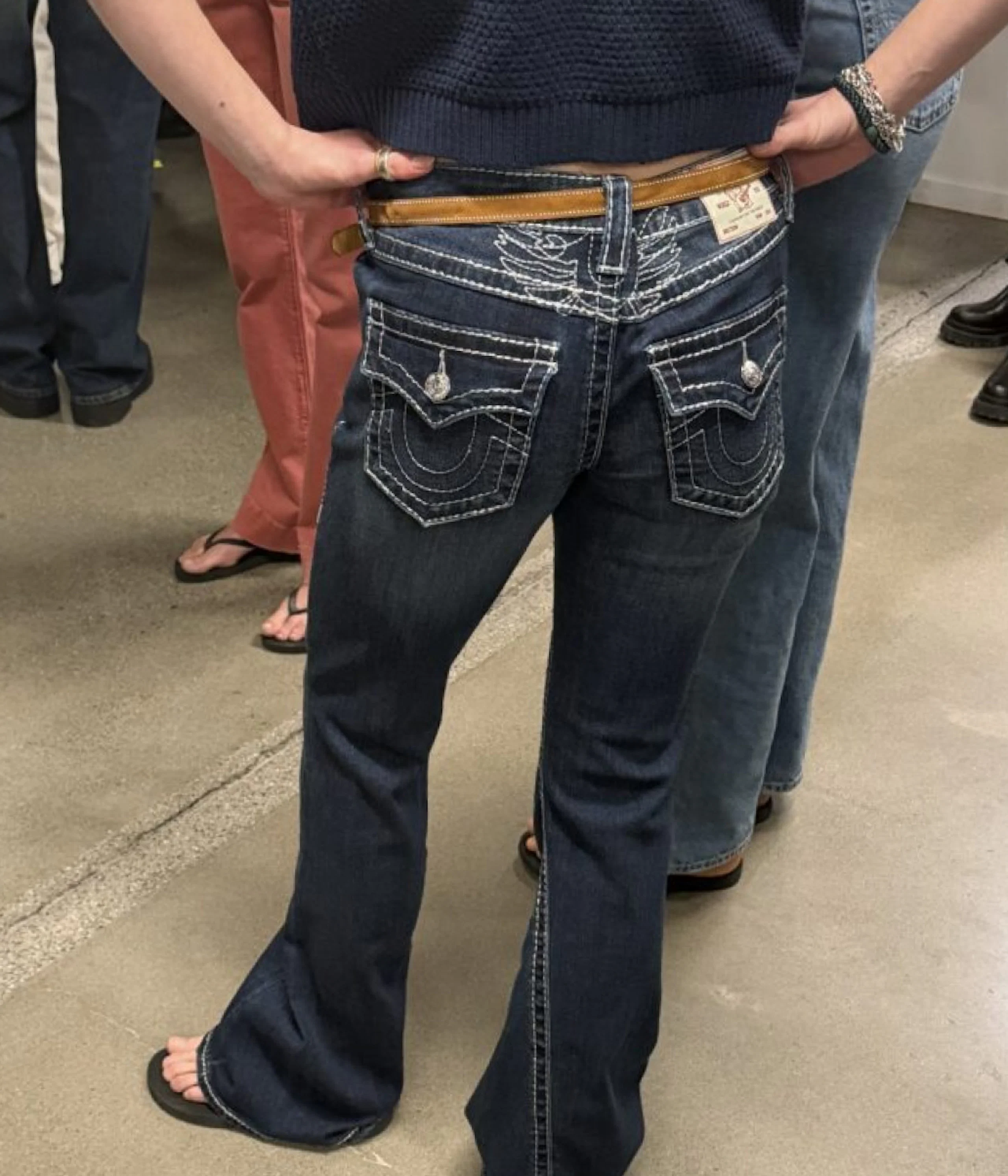 Person sett bakfra iført mørke bootcut-jeans med hvit søm og brunt belte, samt mørk genser.