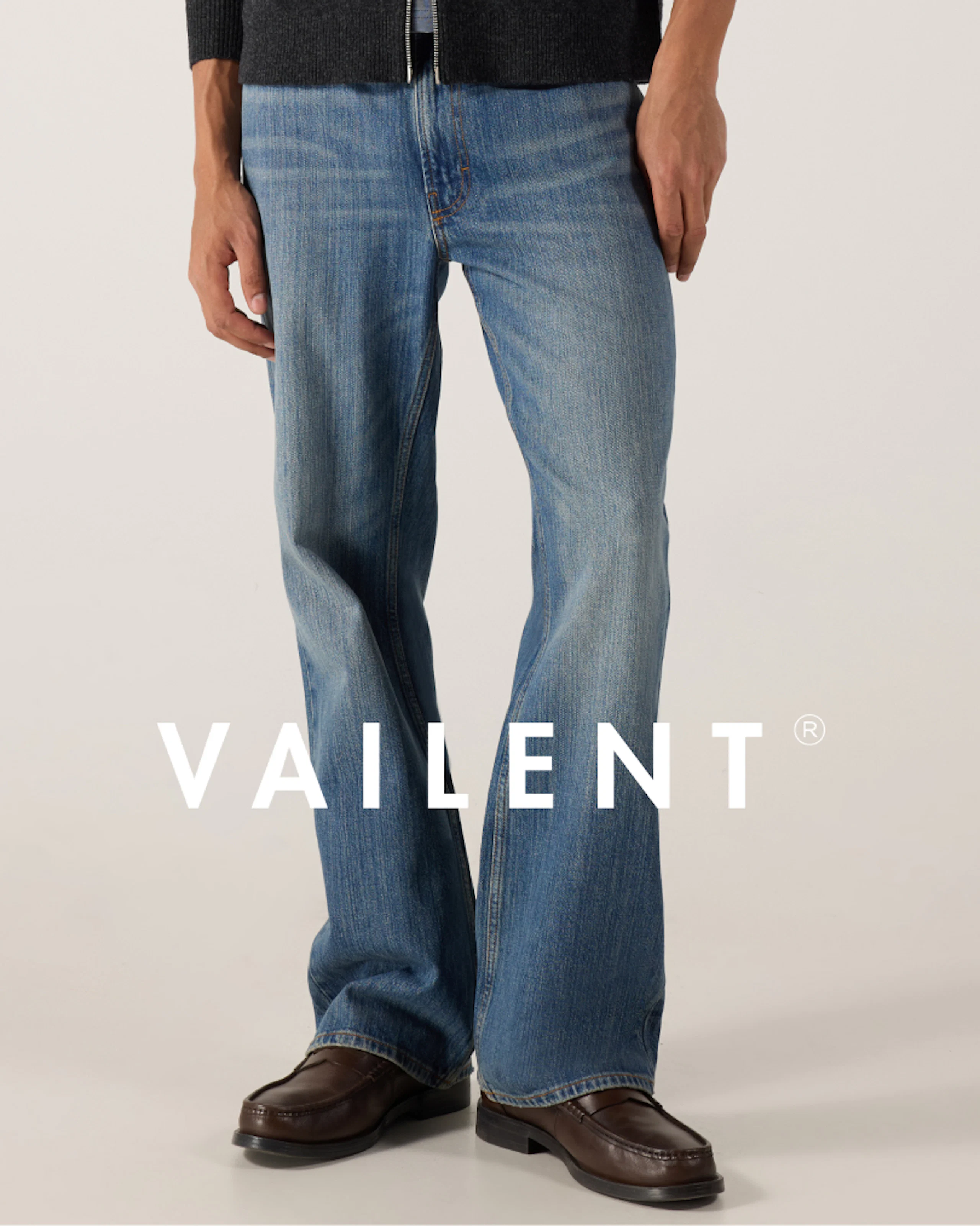 Mann iført vide bukser i medium blå jeansfarge og mørkbrune loafers, kombinert med en grå topp med glidelås. Vailent-logo overlay.