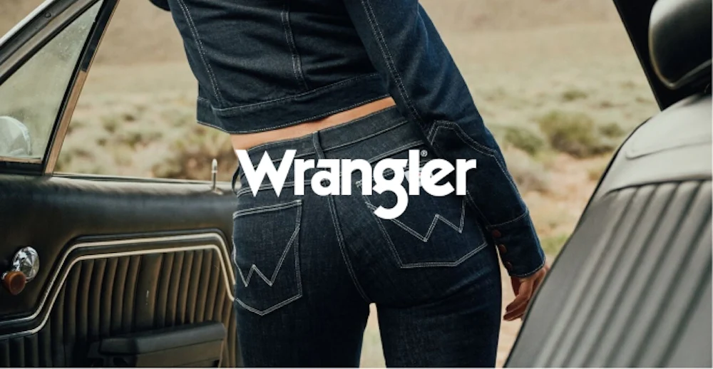 Nainen tummansinisessä Wrangler-farkkutakissa ja -housuissa seisoo vanhan auton vieressä. Wrangler-logo kuvassa.