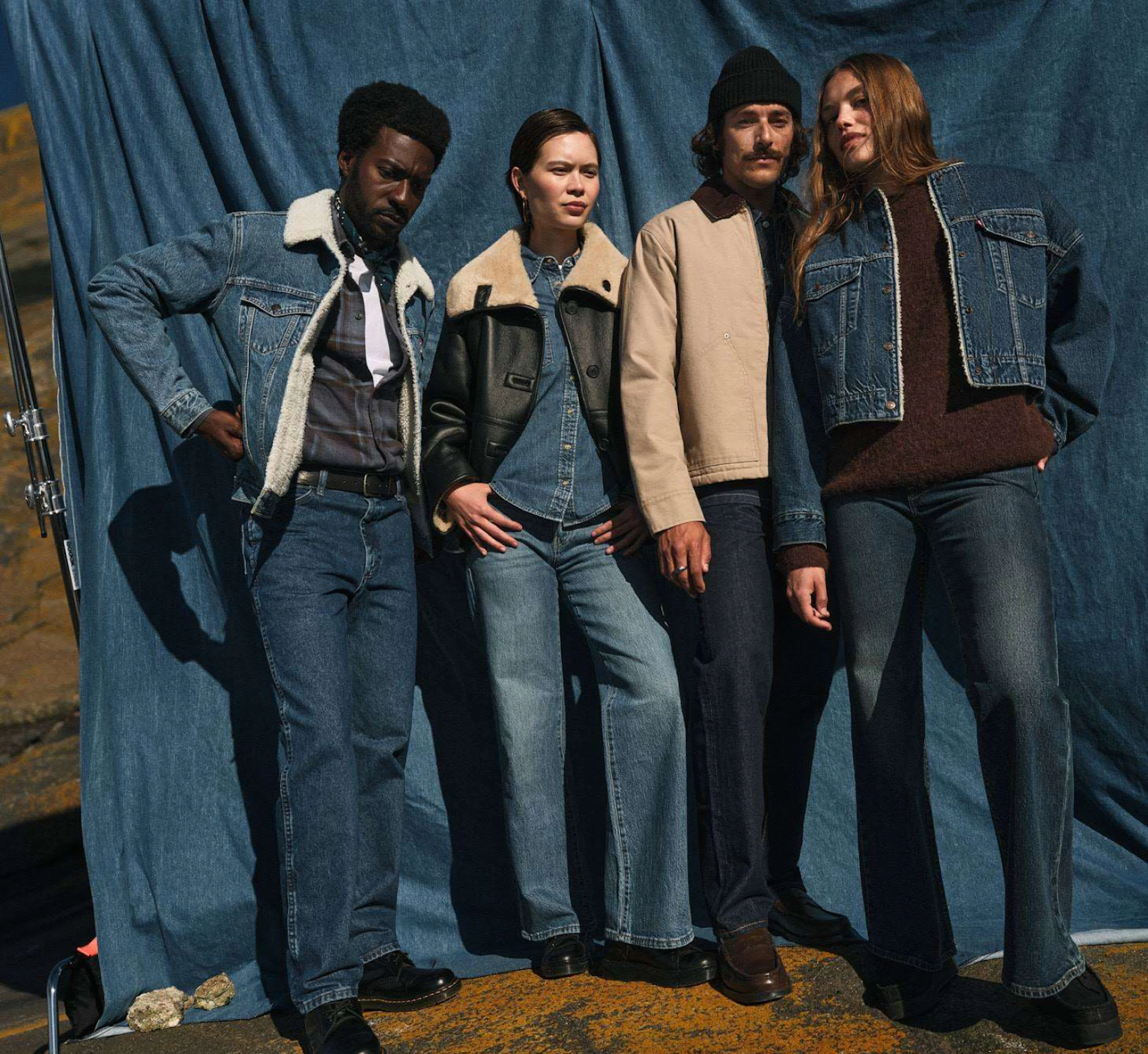 Fyra personer i jeans och höstjackor poserar framför ett blått tyg. Retrostil med denim och shearlingjackor.