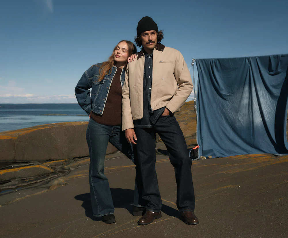 To personer i casual antrekk står utendørs ved havet. Hun har denimjakke, han beige jakke og svart lue.