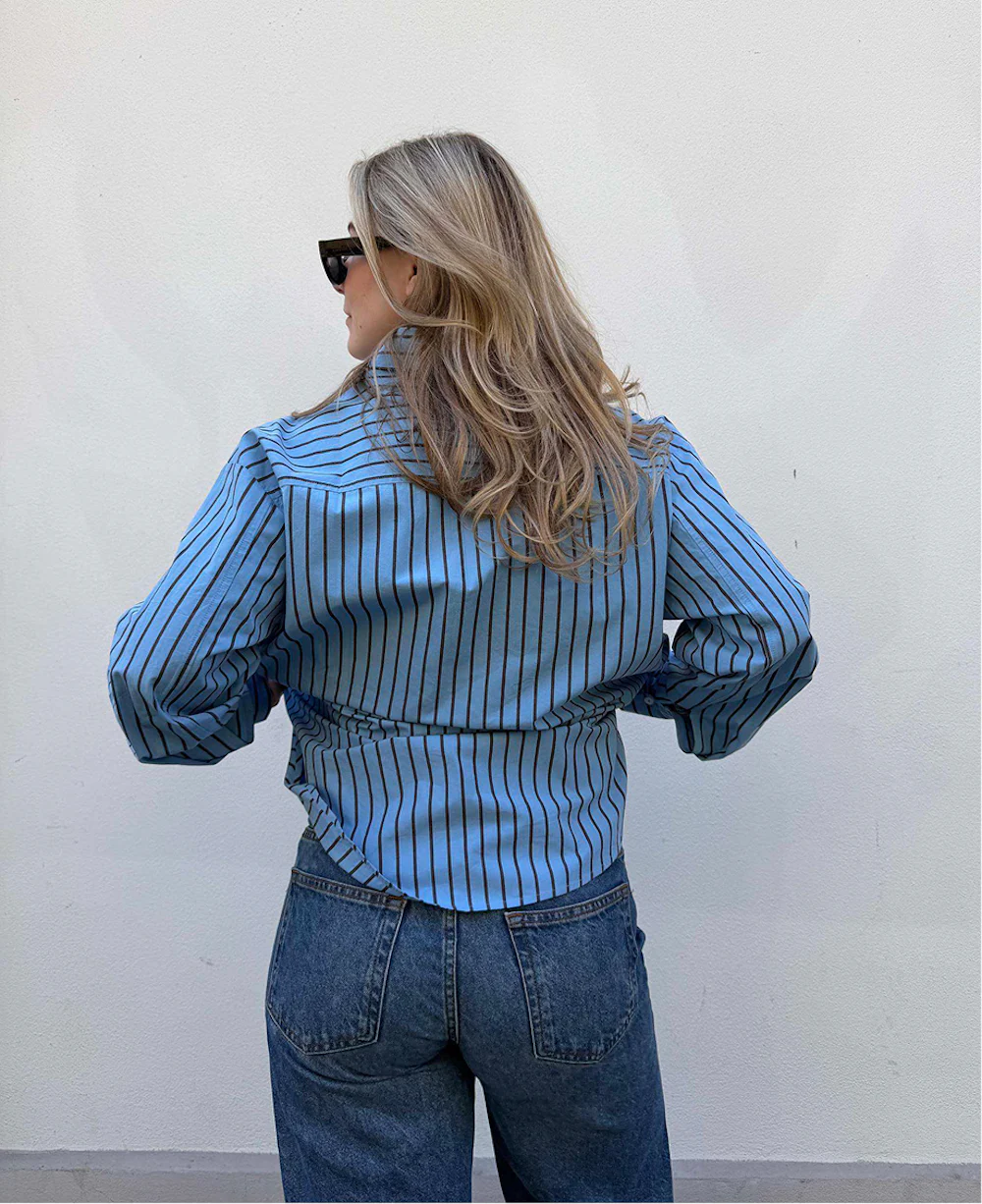 Kvinne sett bakfra med blå og svart stripete skjorte, blå jeans og solbriller, langt blondt hår.
