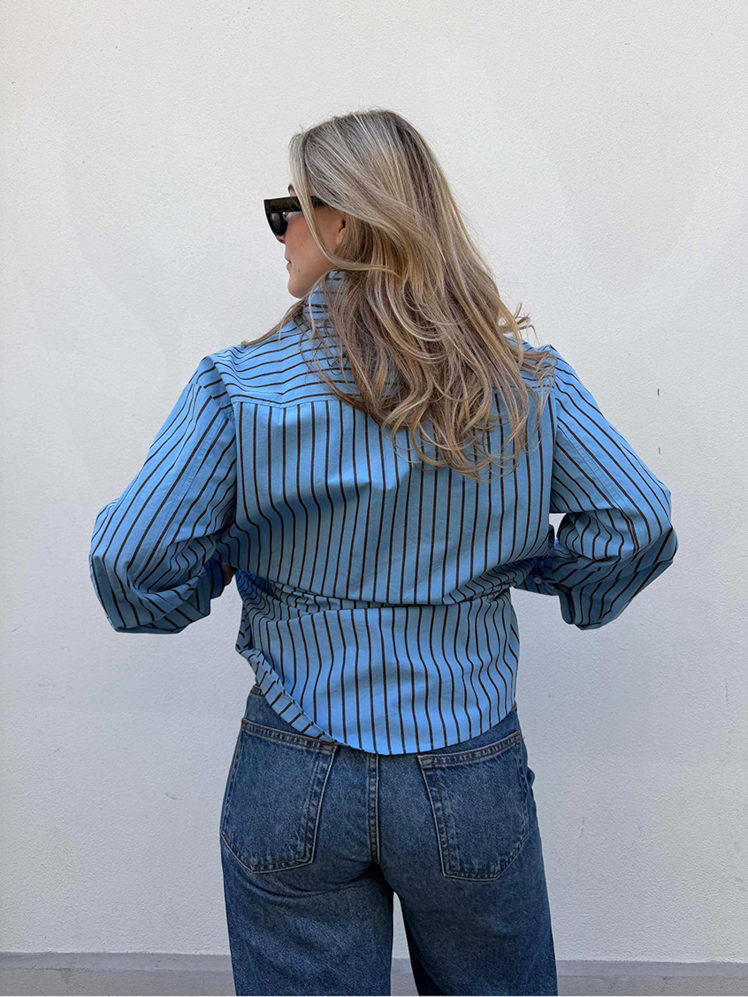 Kvinne sett bakfra med blå og svart stripete skjorte, blå jeans og solbriller, langt blondt hår.