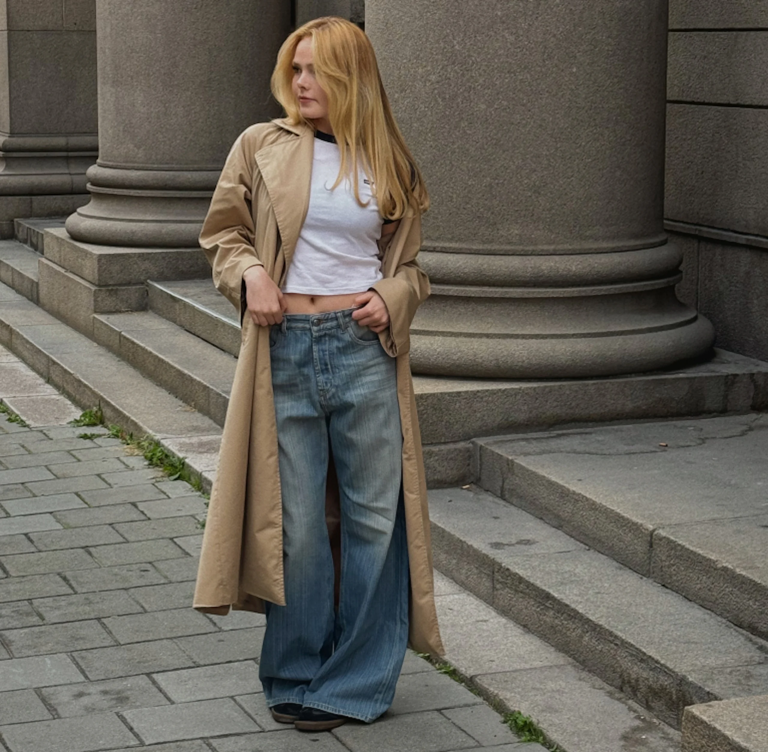 Nuori nainen seisoo kivipylväiden edessä beige trenssitakki, valkoinen crop top ja leveälahkeiset farkut yllään.
