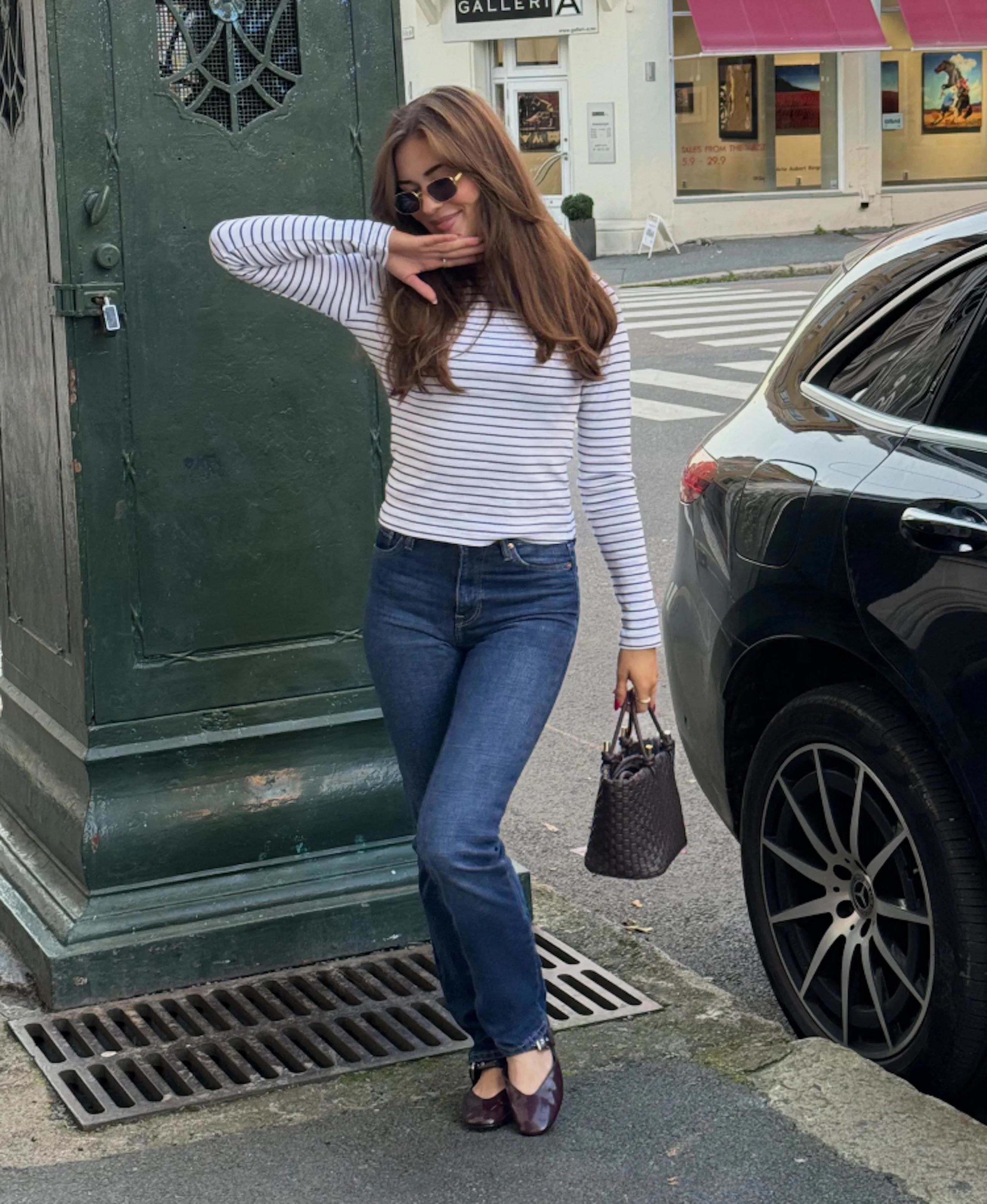 Ung kvinna i randig topp, blå jeans och solglasögon poserar på gatan med en mörk väska bredvid en svart bil.