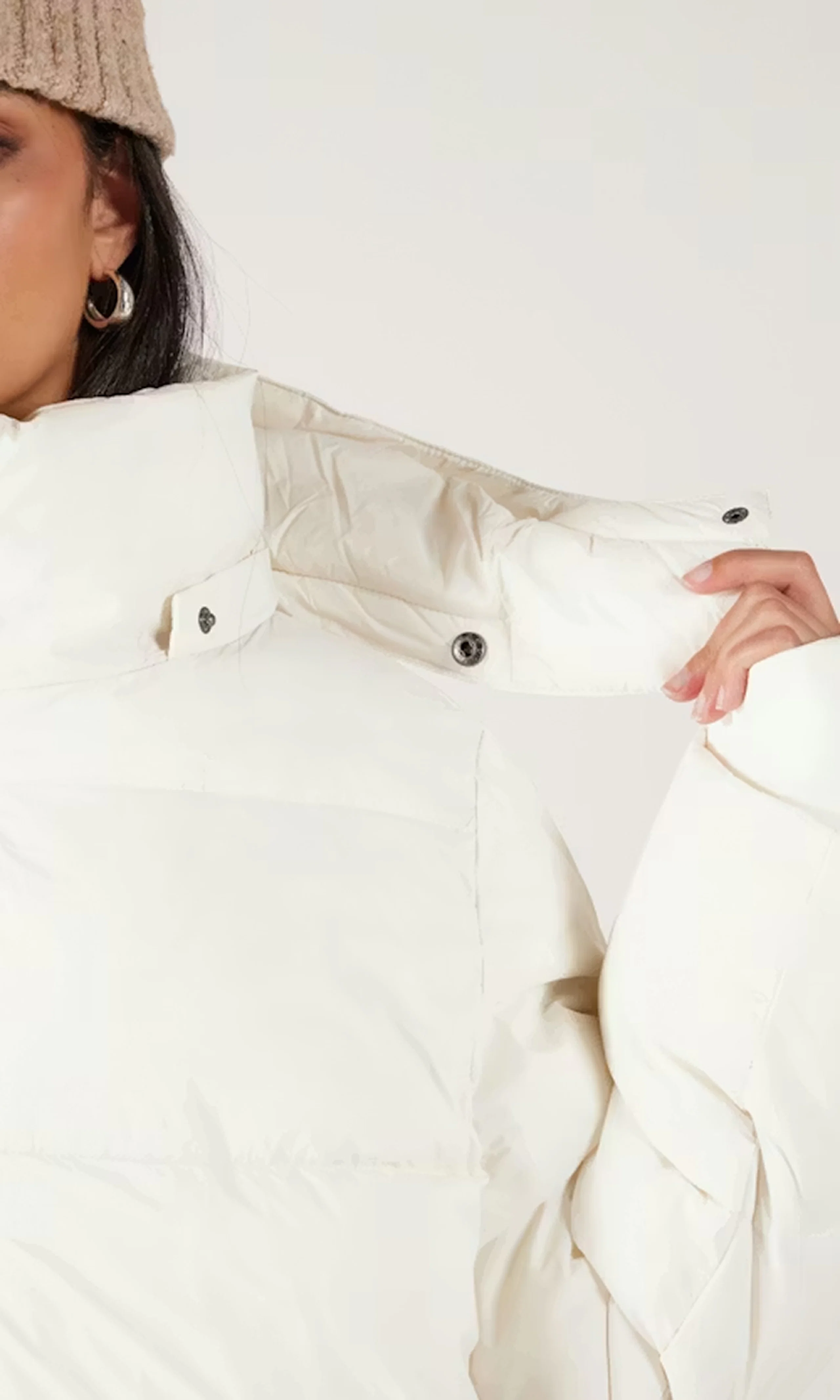 Woman wearing a cream puffer jacket and beige knit beanie, holding the jacket's padded collar with her hand, cropped to show hoop earring and upper torso.