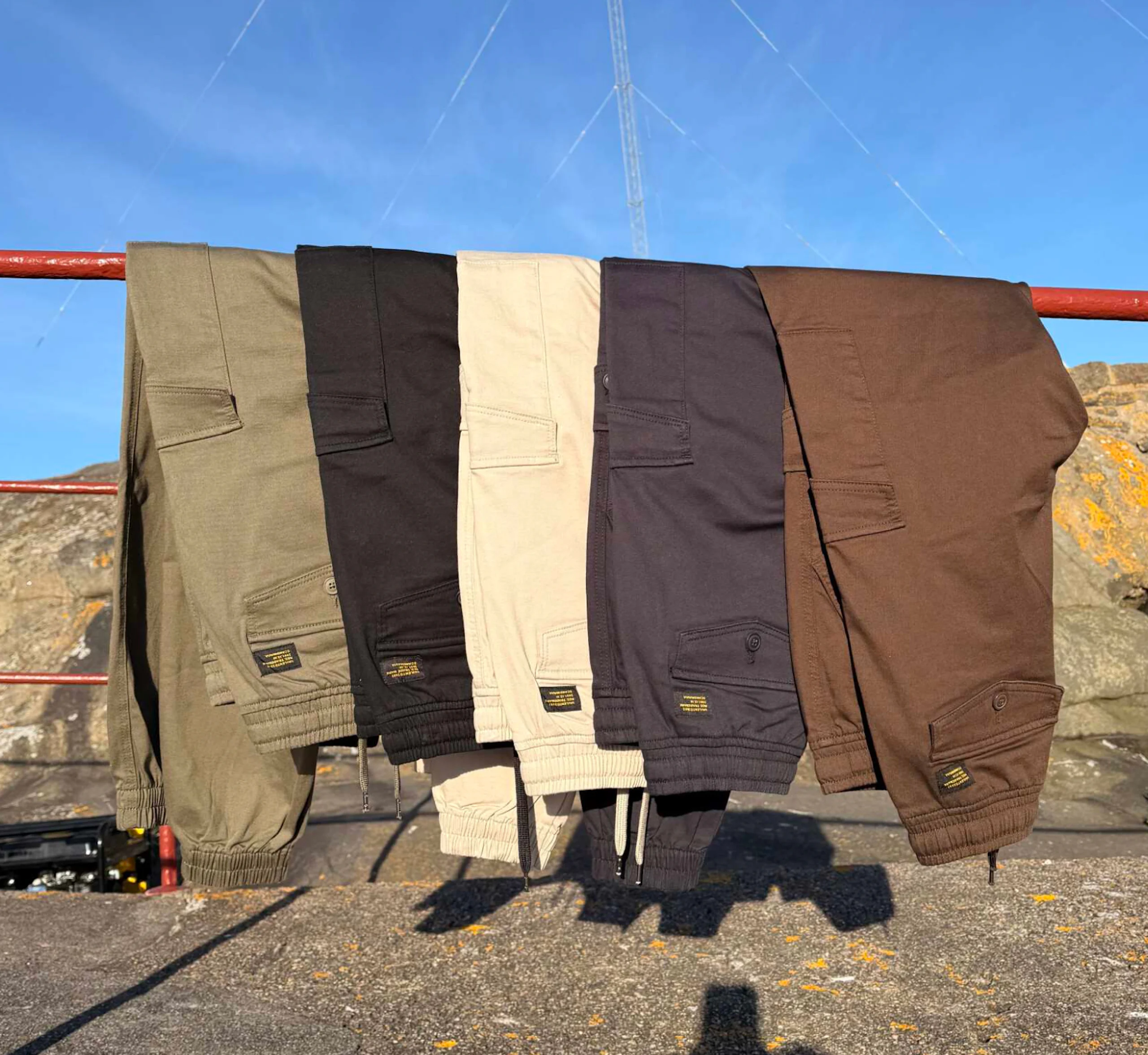 Fem cargobyxor i olika färger hänger på ett rött räcke utomhus mot blå himmel. Färgerna är grön, svart, beige, mörkgrå och brun.