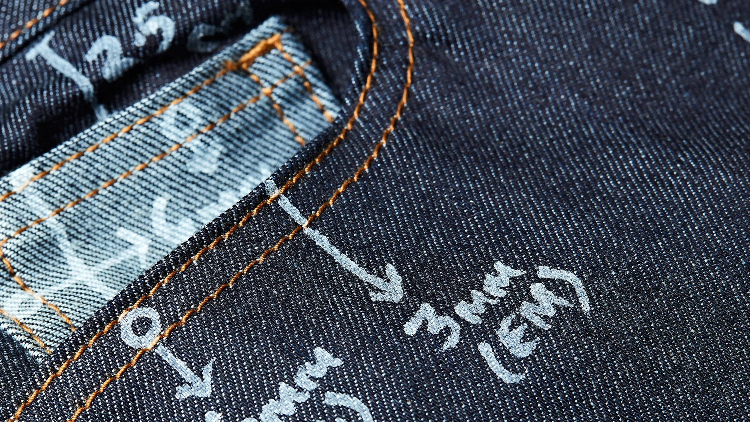 Nærbilde av hjørnet på en mørk denimlomme med oransje overlock og hvite trykte størrelsesmarkeringer på teksturert stoff.