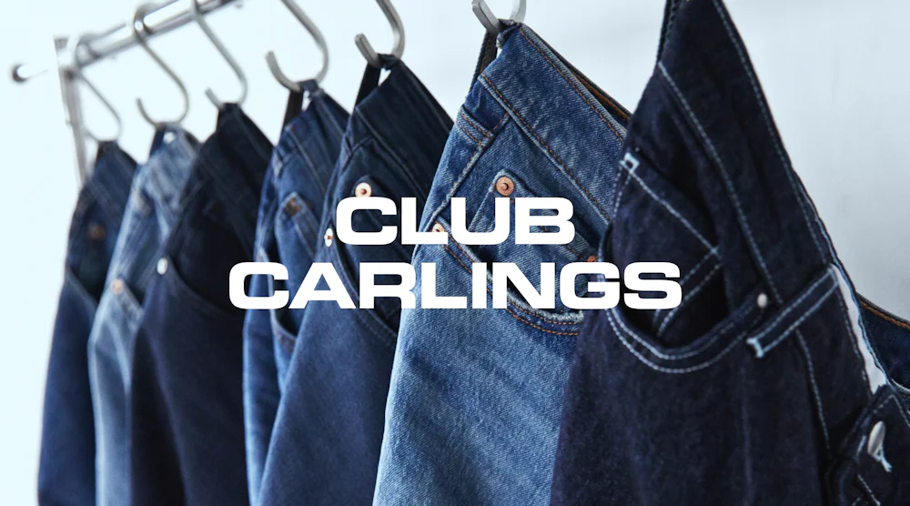 Flere blå jeansbukser hengt på metallkroker, med teksten "Club Carlings" i hvitt i forgrunnen.