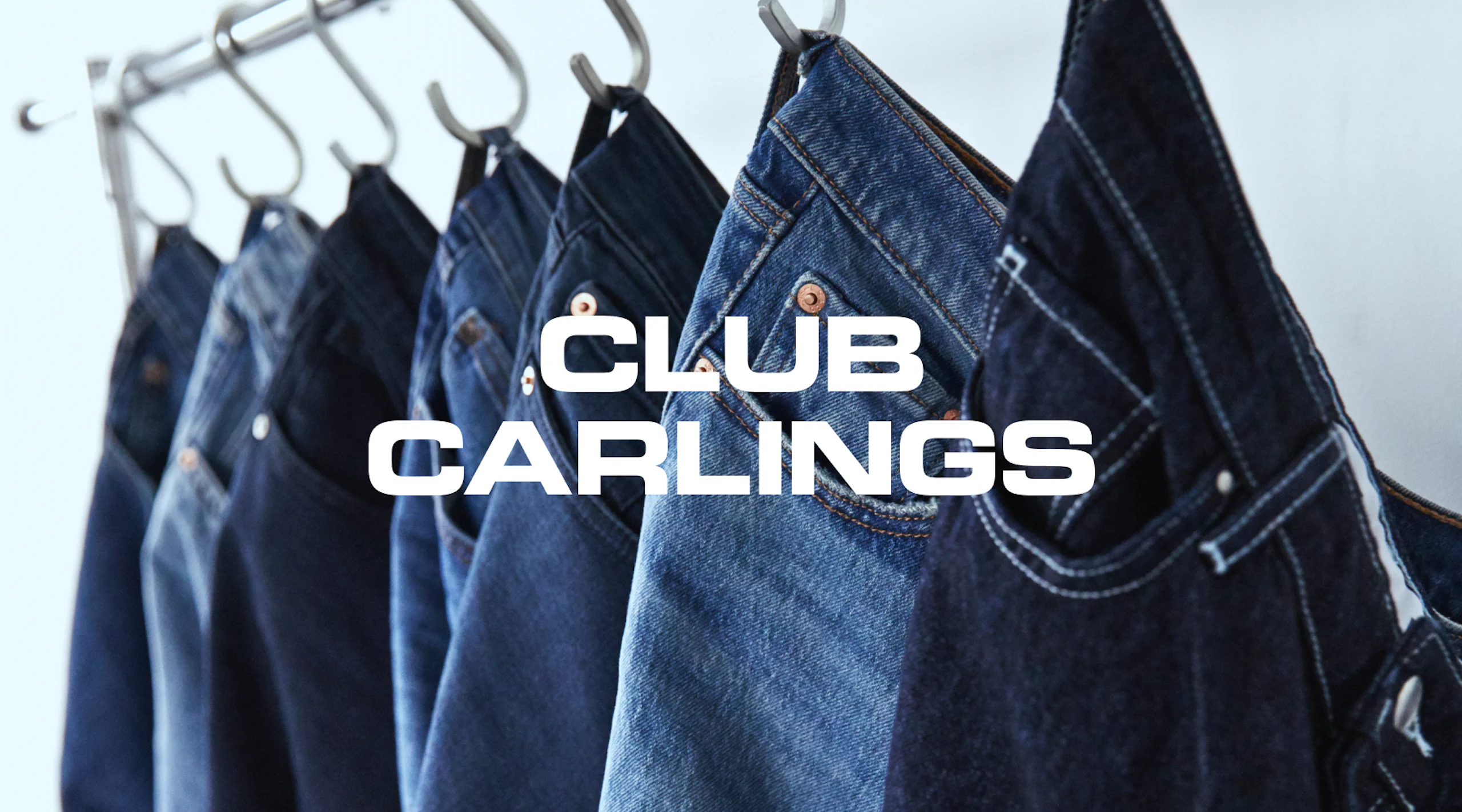 Useita sinisiä farkkuja roikkuu metallisilla koukuilla rekkiin. Teksti "Club Carlings" näkyy kuvassa.