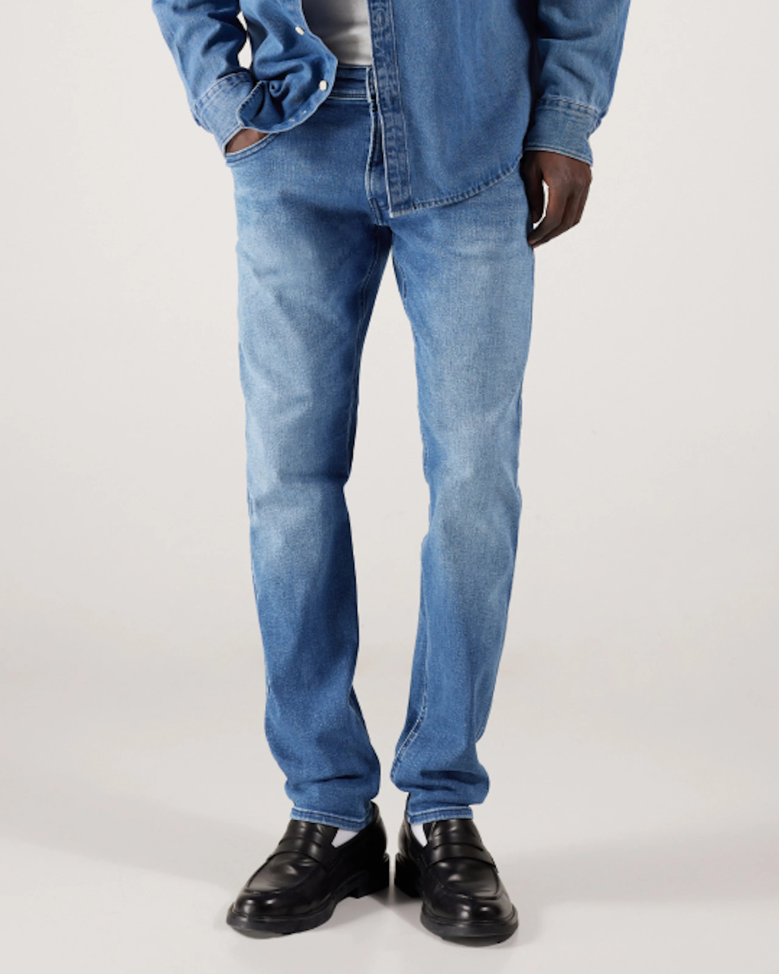 Man som bär mellanblå slim-fit jeans, en jeansskjorta och svarta loafers, visad från midjan och ned mot en ljusgrå bakgrund.