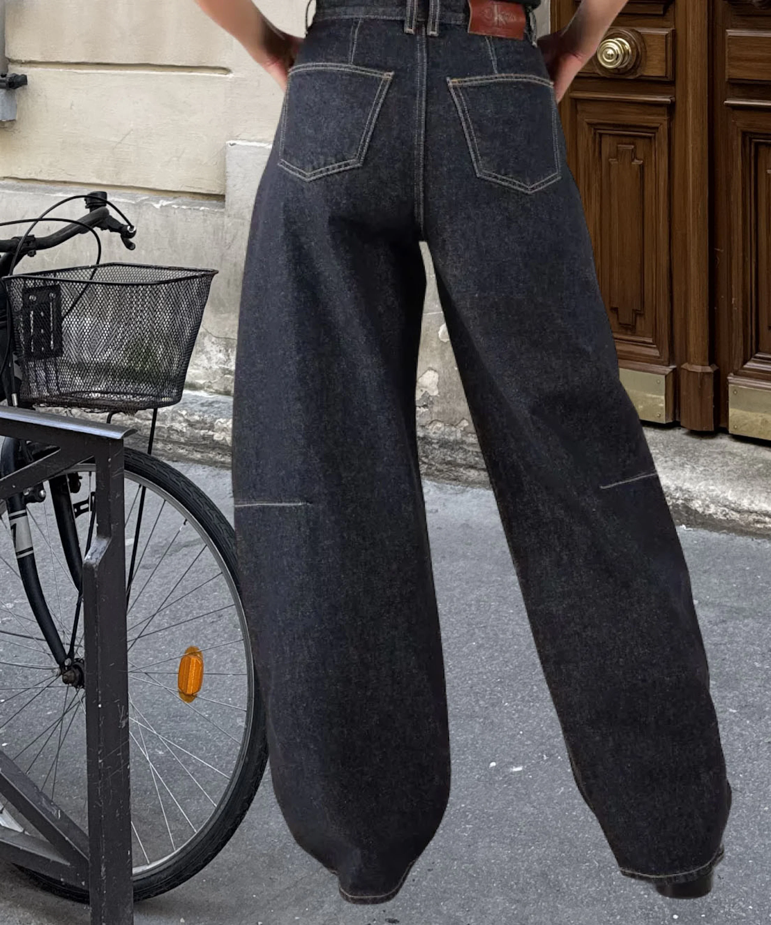 Person sett bakfra iført mørke wide-leg jeans med synlige sømmer og lærbelte, stående på en gate ved en sykkel.